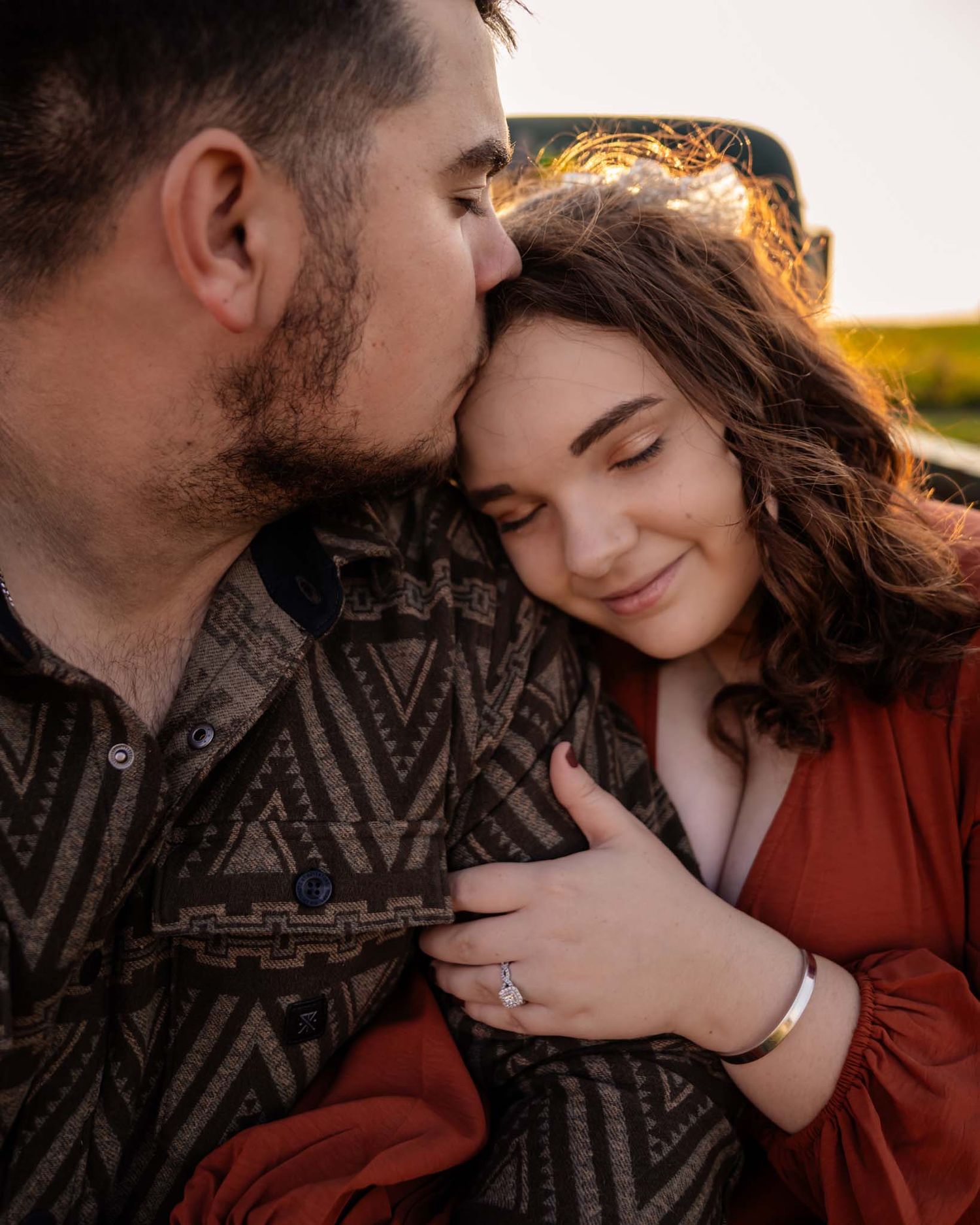 A man kisses a smiling woman on the head while embracing her outdoors during golden hour.