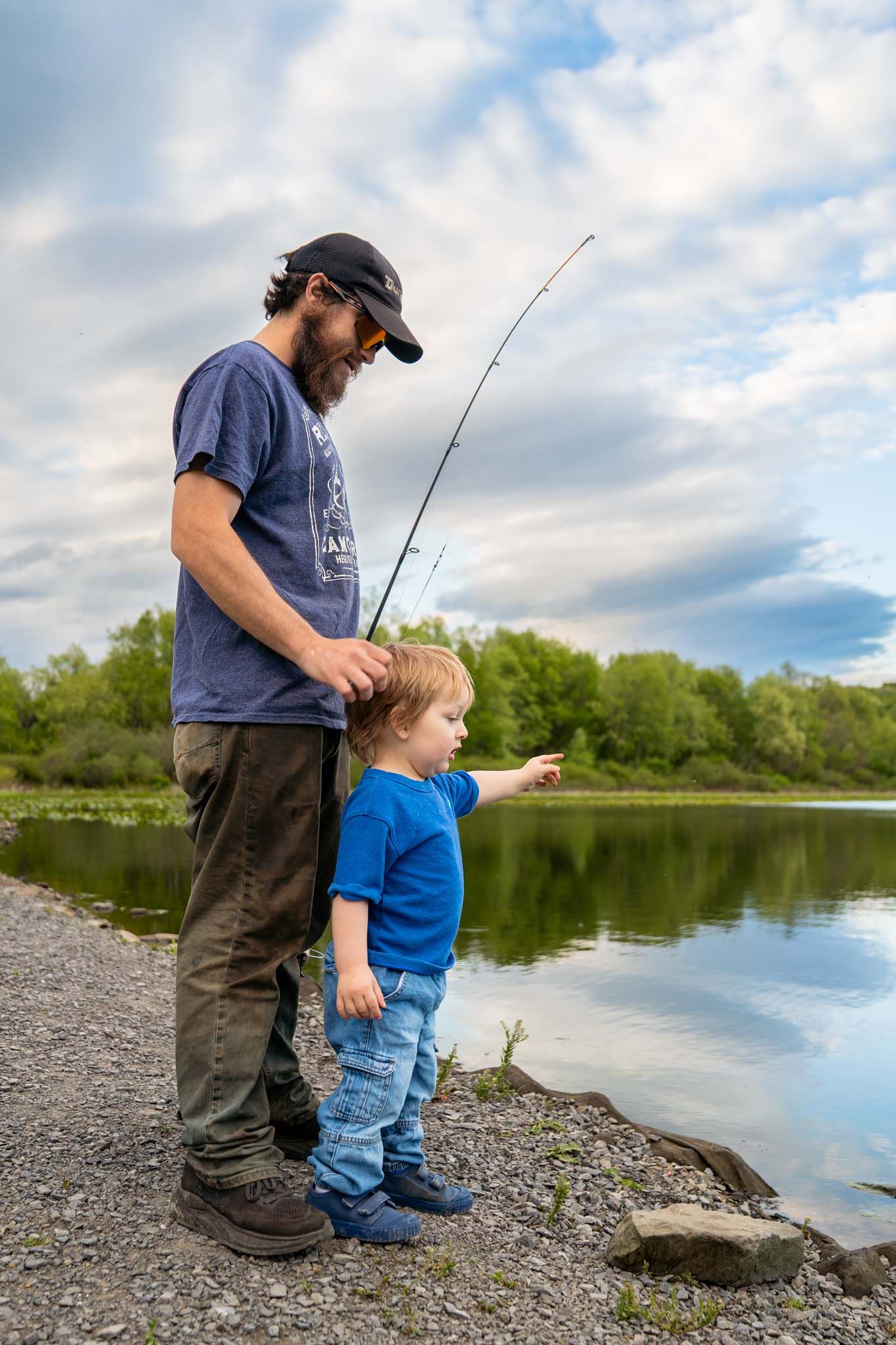 Father and toddler son standing by lake shore as child points while dad holds fishing rod.