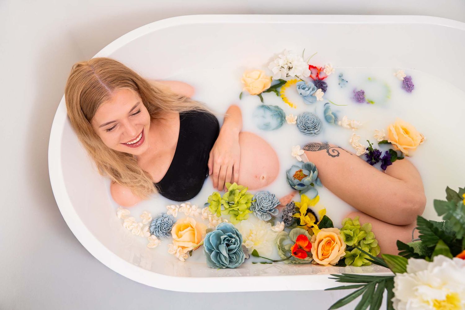 Smiling pregnant woman relaxes in floral milk bath during creative maternity photo session.