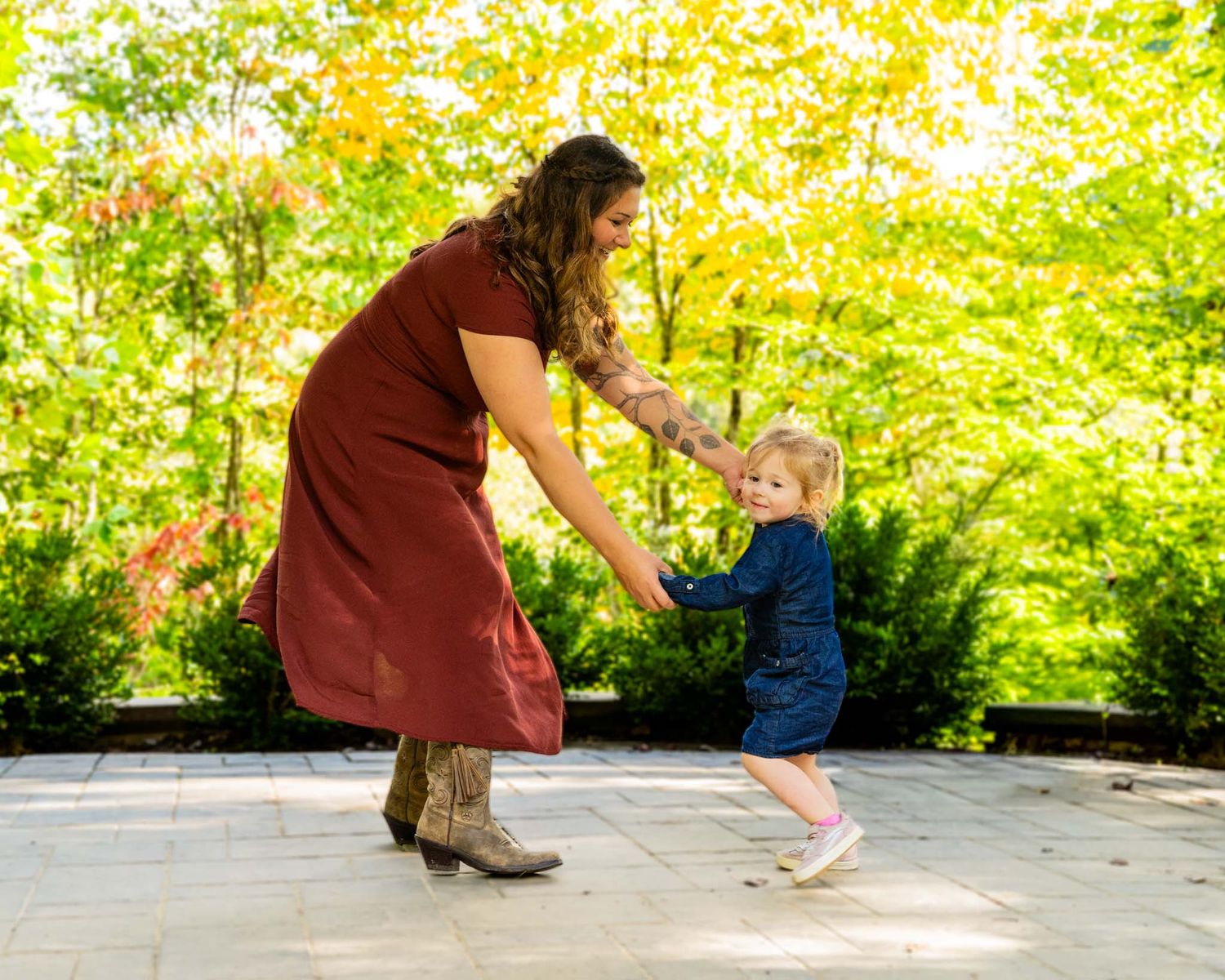 A mother in a red dress and cowboy boots dances playfully with her toddler on a stone patio surrounded by autumn foliage.
