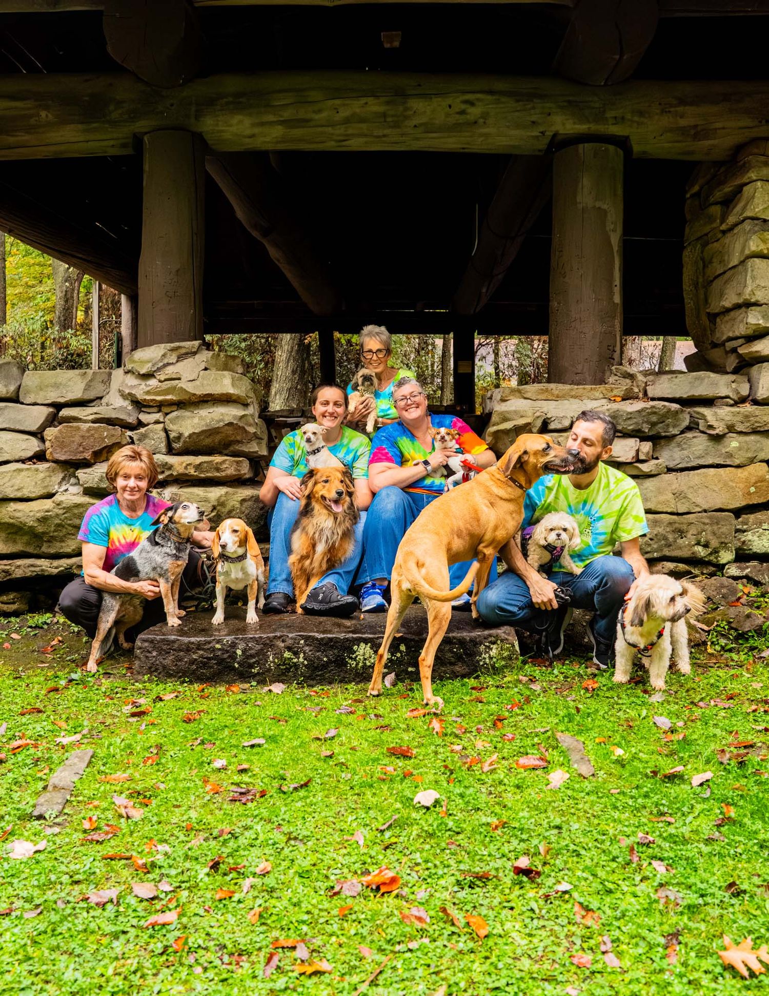Group of people in tie-dye shirts posing outdoors with multiple dogs near a rustic stone structure.