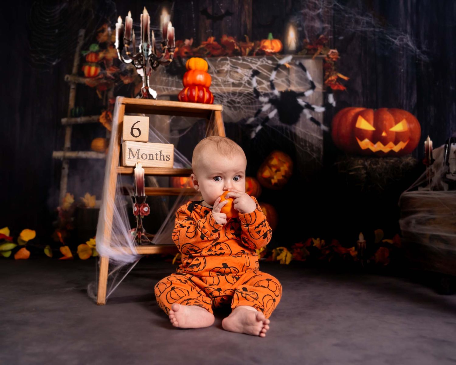 Baby in orange Halloween outfit sits with pumpkins and spiderweb decor, marking 6 months milestone.