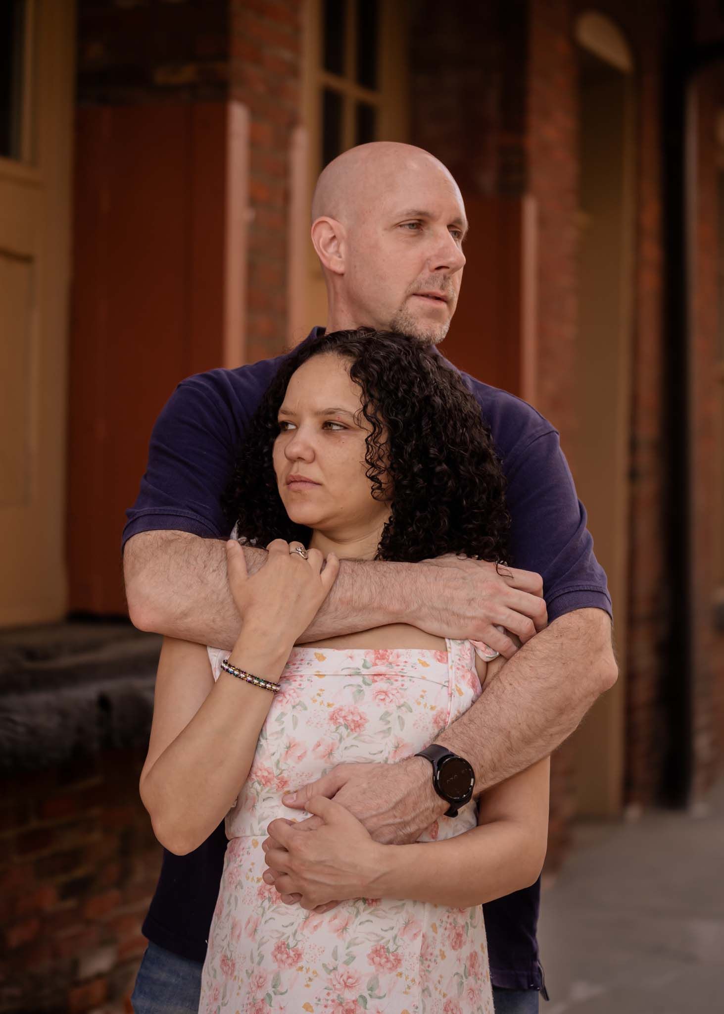 Bald man tenderly embracing woman with curly hair in front of a rustic brick building outdoors.