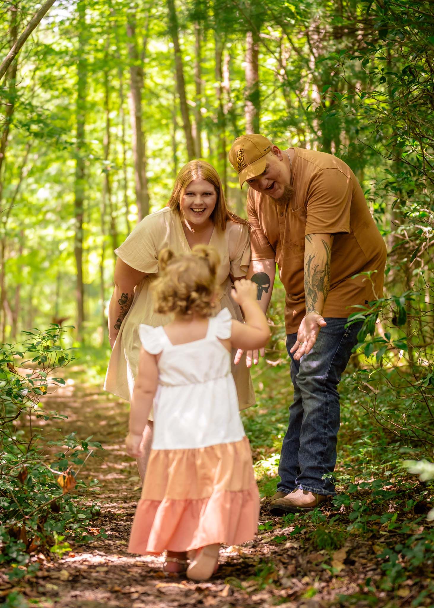 A joyful family moment on a forest path as a toddler girl in a white and peach dress runs toward her smiling parents.