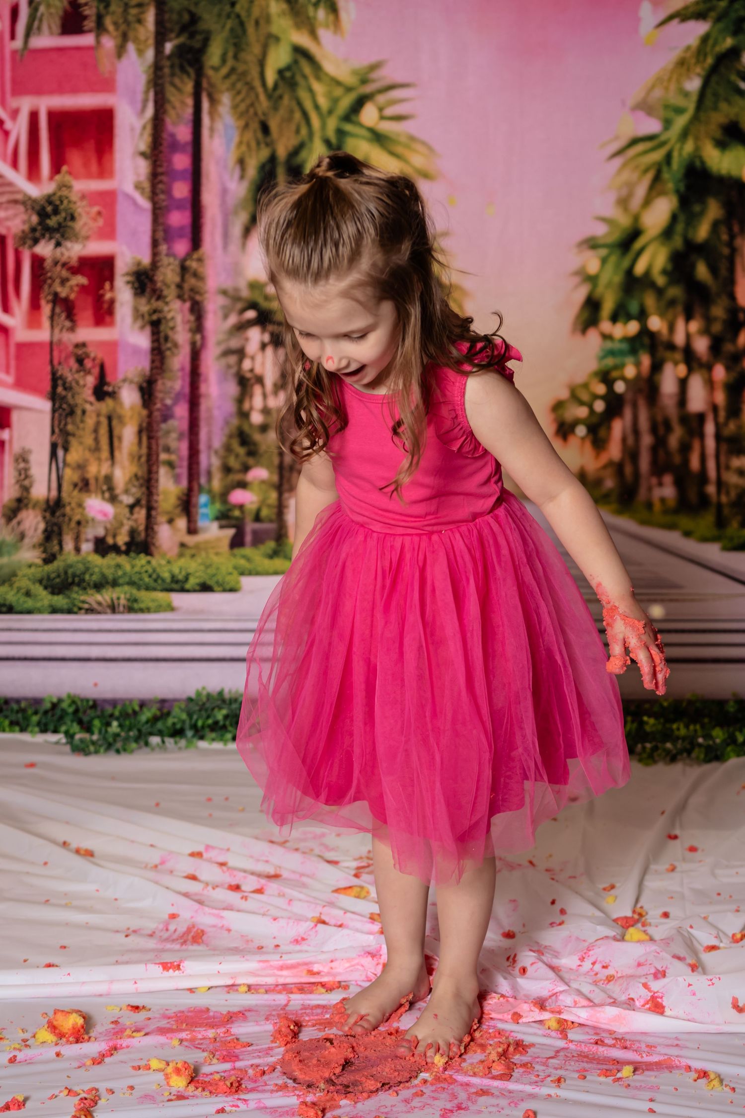A young girl in a pink tutu dress smiles while standing barefoot on a cake-smashed white surface at a birthday party.