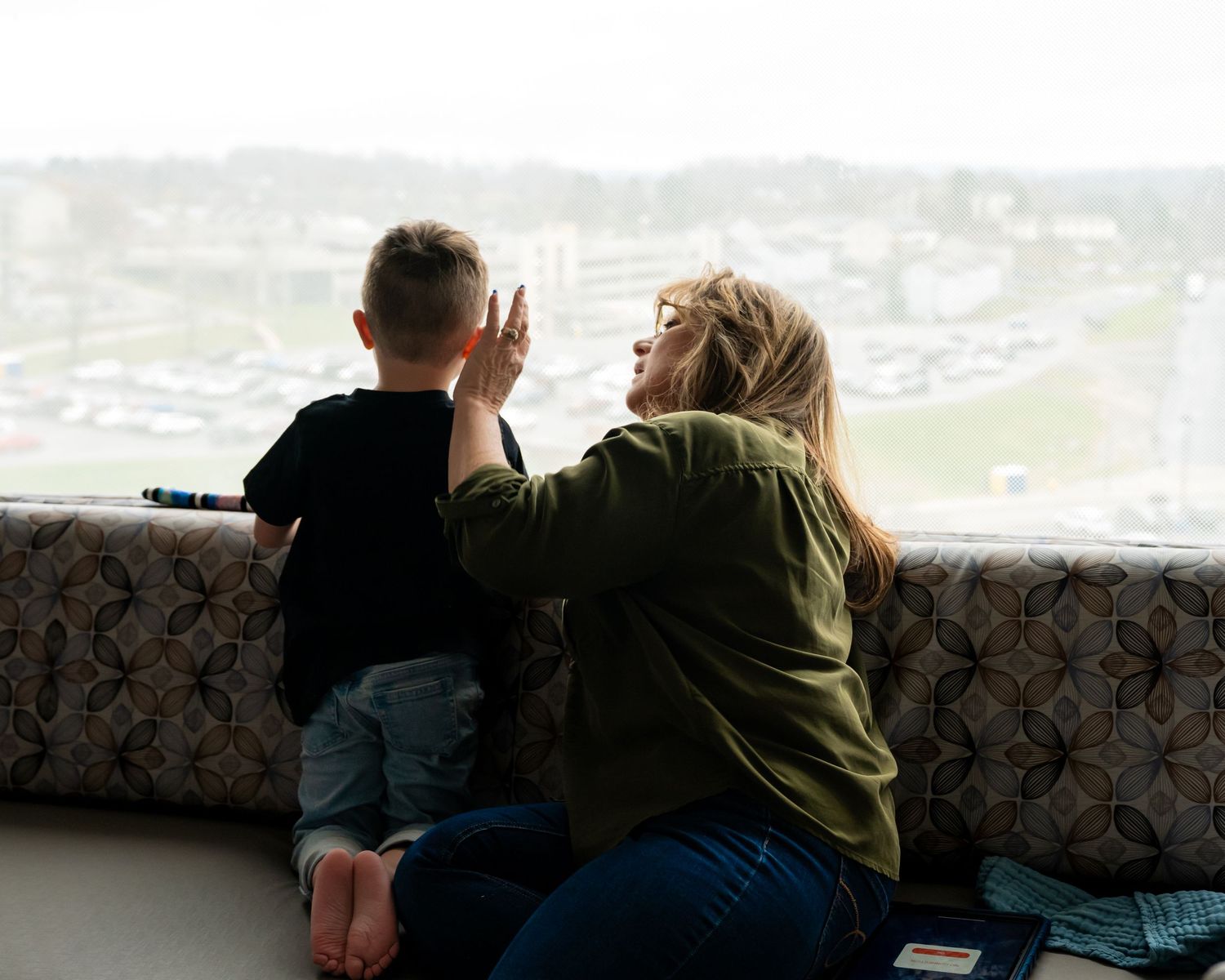 A mother and young boy share a high-five moment by a large window overlooking a cityscape.