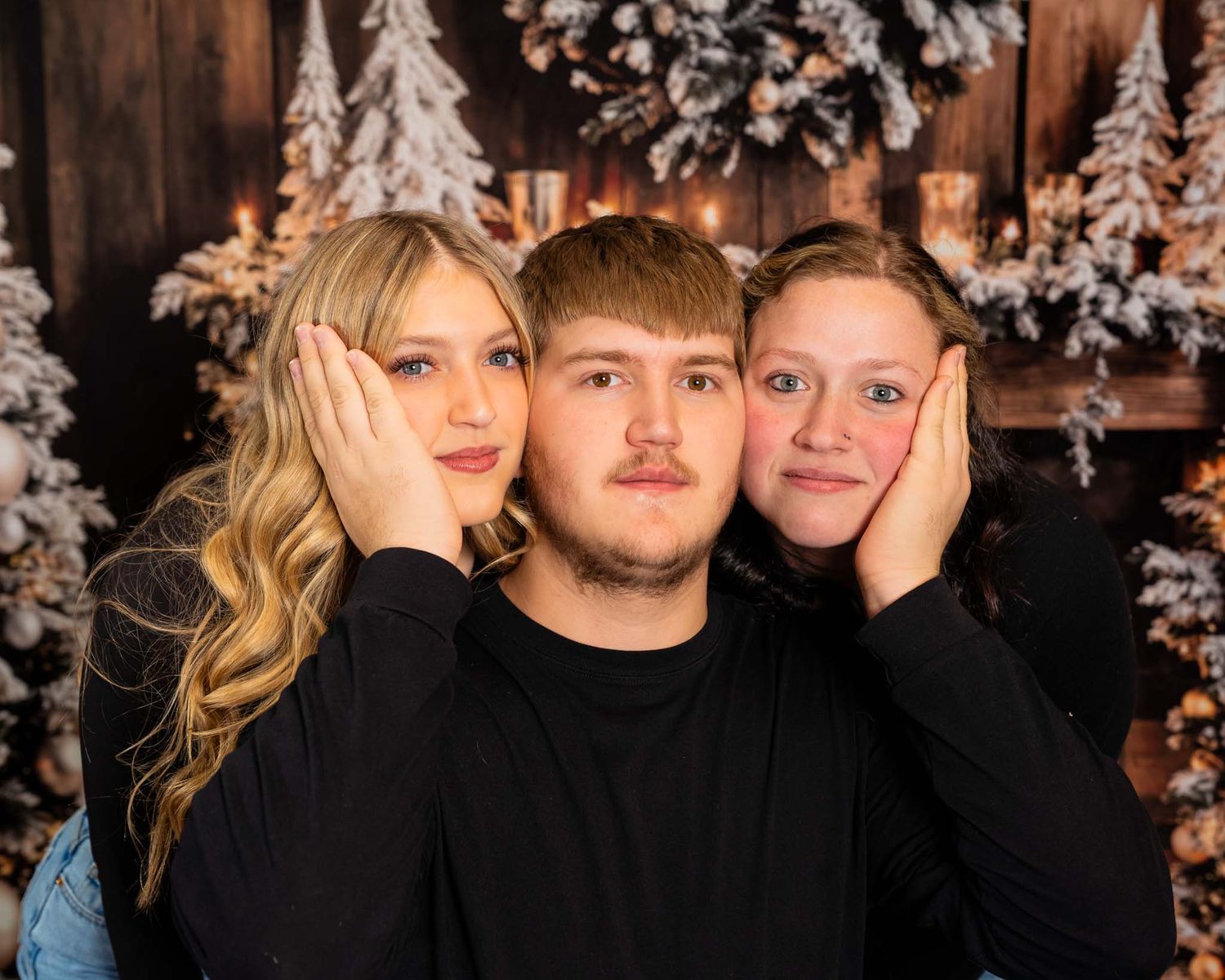 Three teenagers pose for a festive holiday portrait in front of a snowy Christmas tree backdrop in matching black outfits.