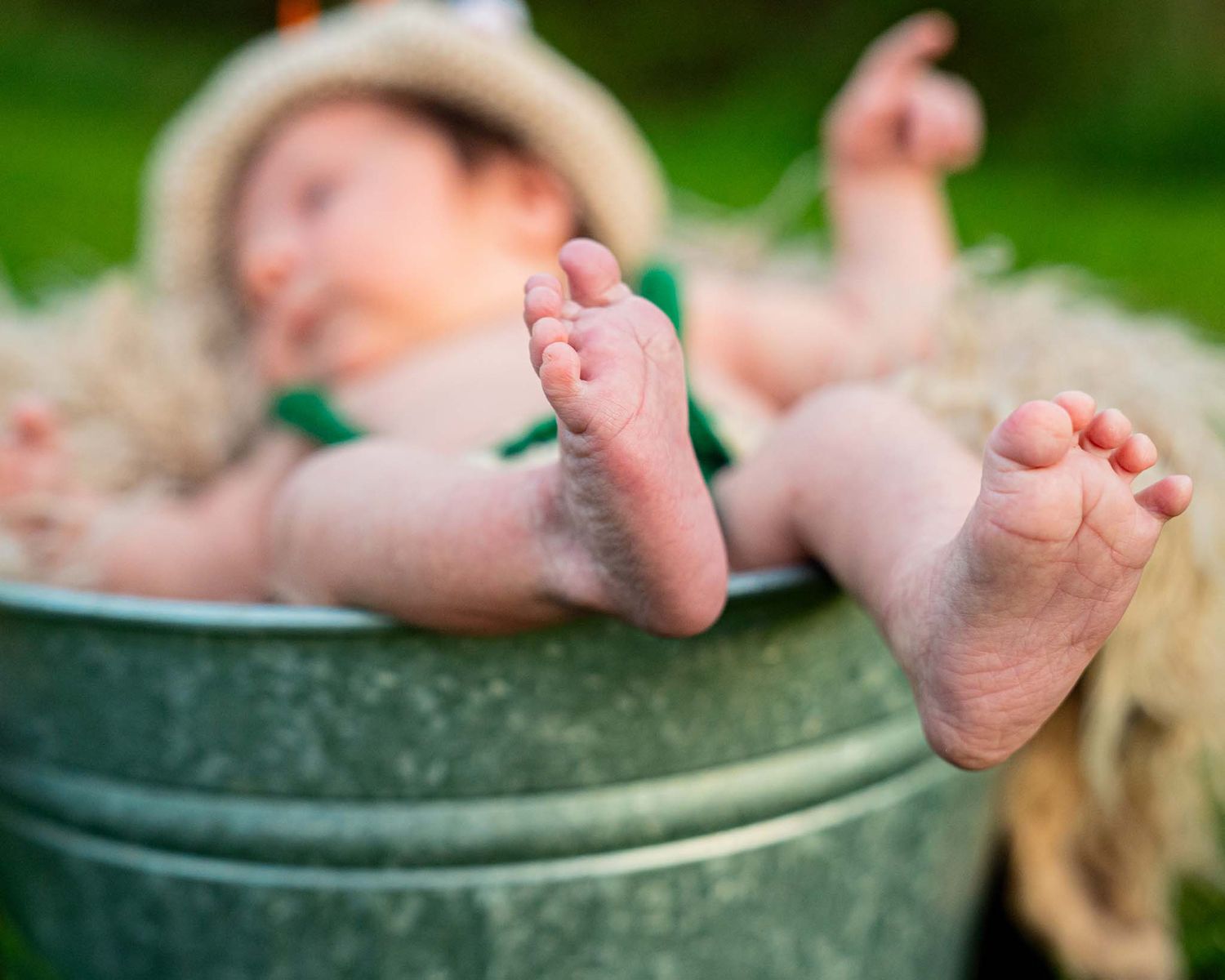 Adorable newborn baby with tiny feet propped up in a rustic metal bucket filled with hay outdoors.