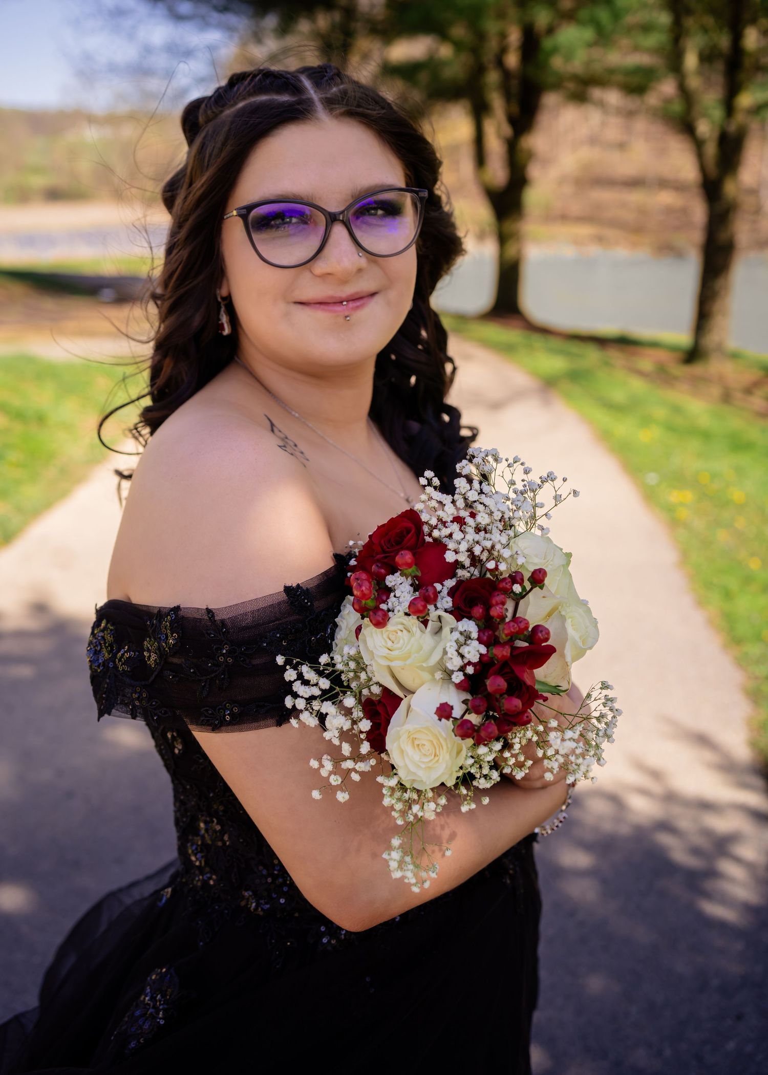 Young woman wearing glasses in a black off-shoulder gown holding a bouquet of red and white flowers.