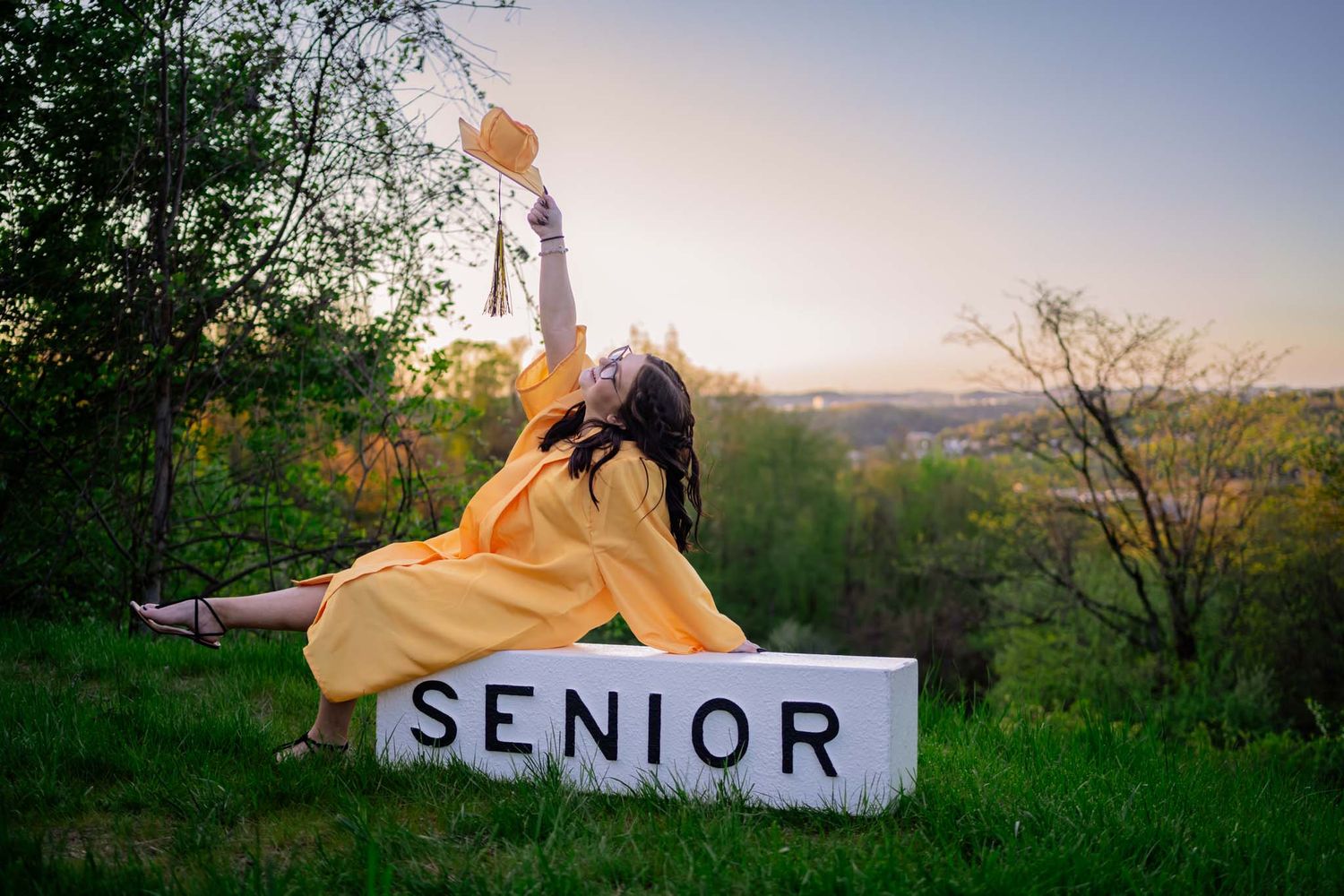Female senior in gold graduation gown joyfully posing on a white Senior sign block at sunset.
