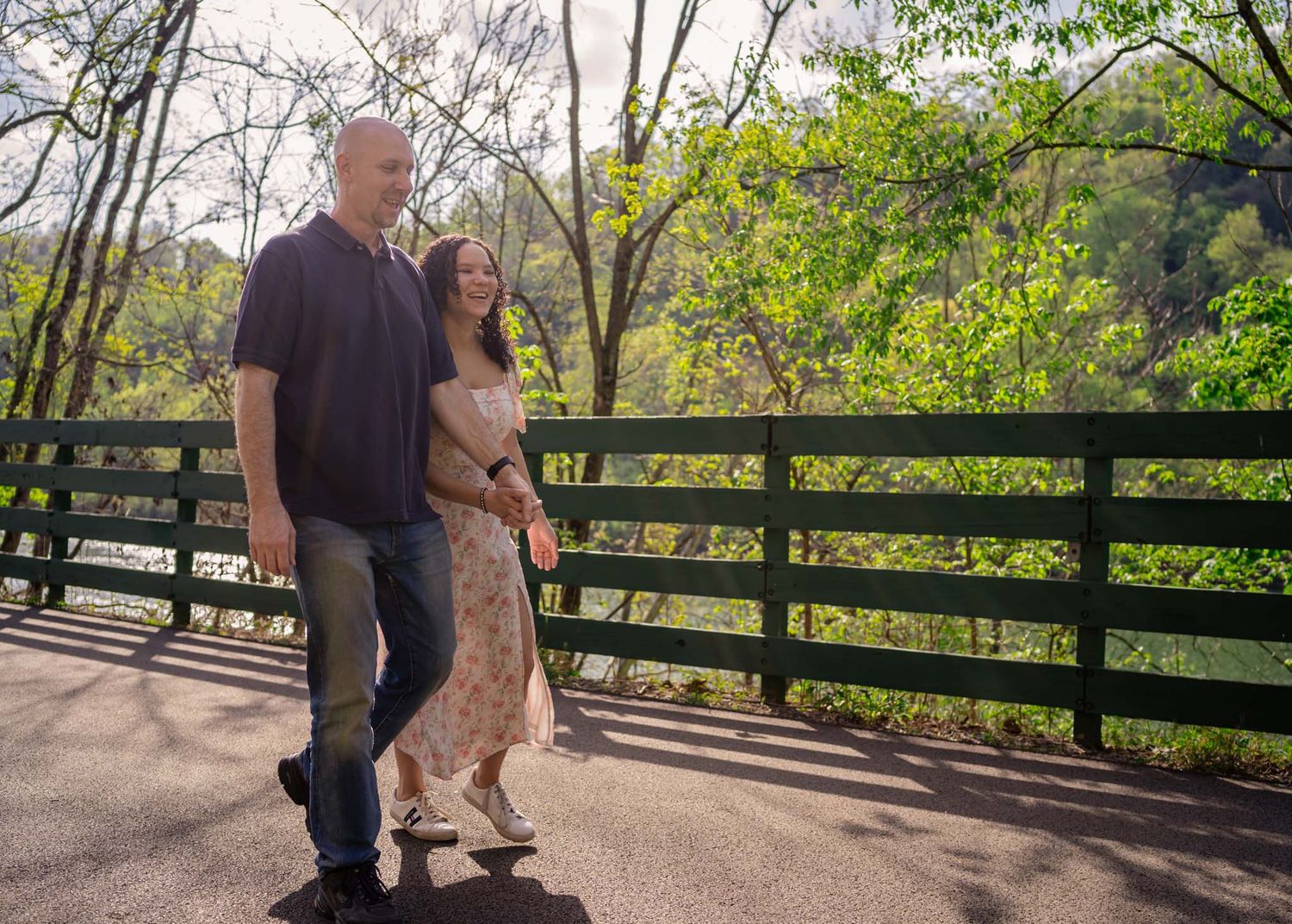 Couple walking hand in hand on a sunny bridge path surrounded by vibrant green spring trees.