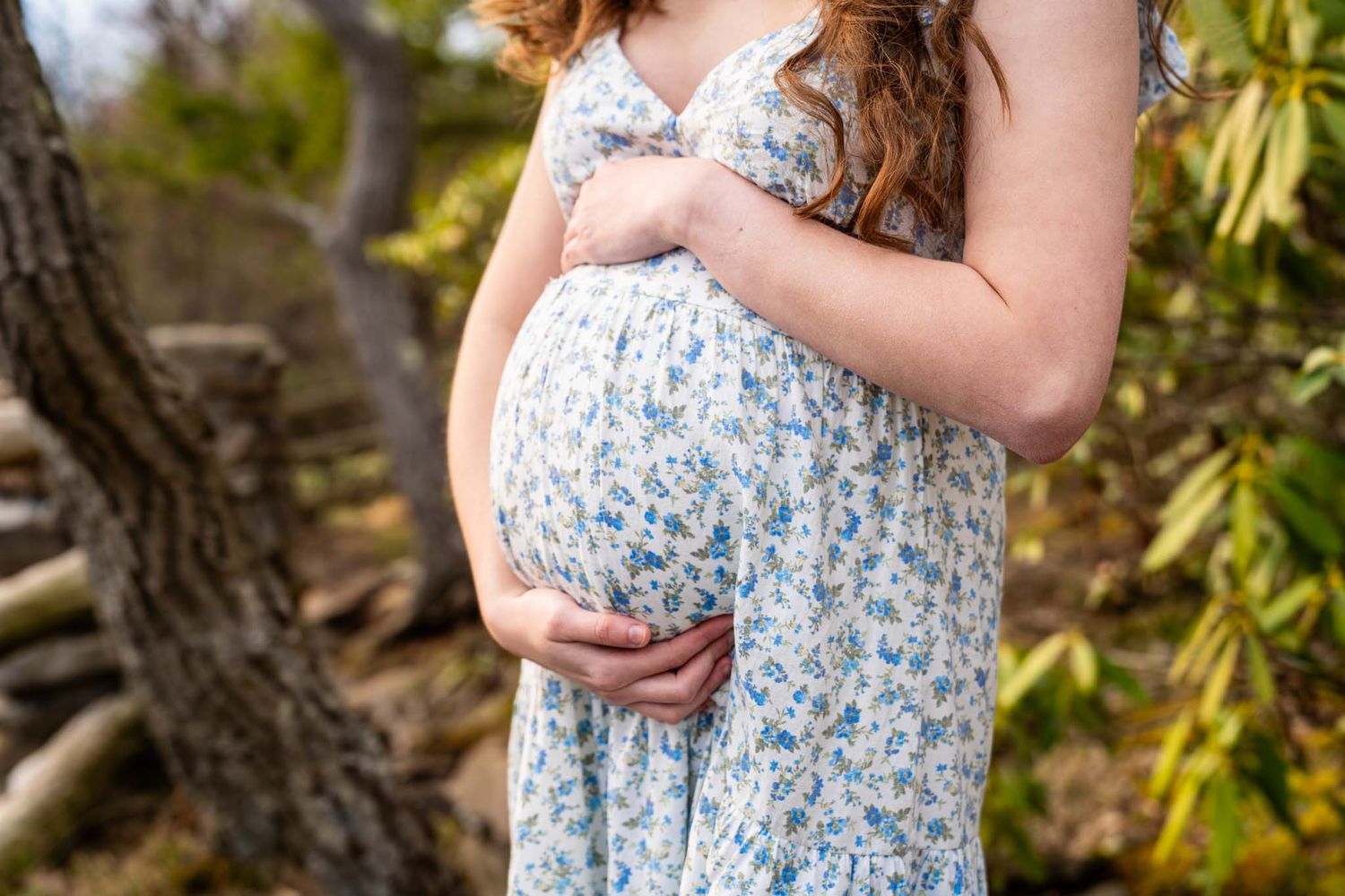 Pregnant woman in floral sundress cradles baby bump outdoors among lush greenery.