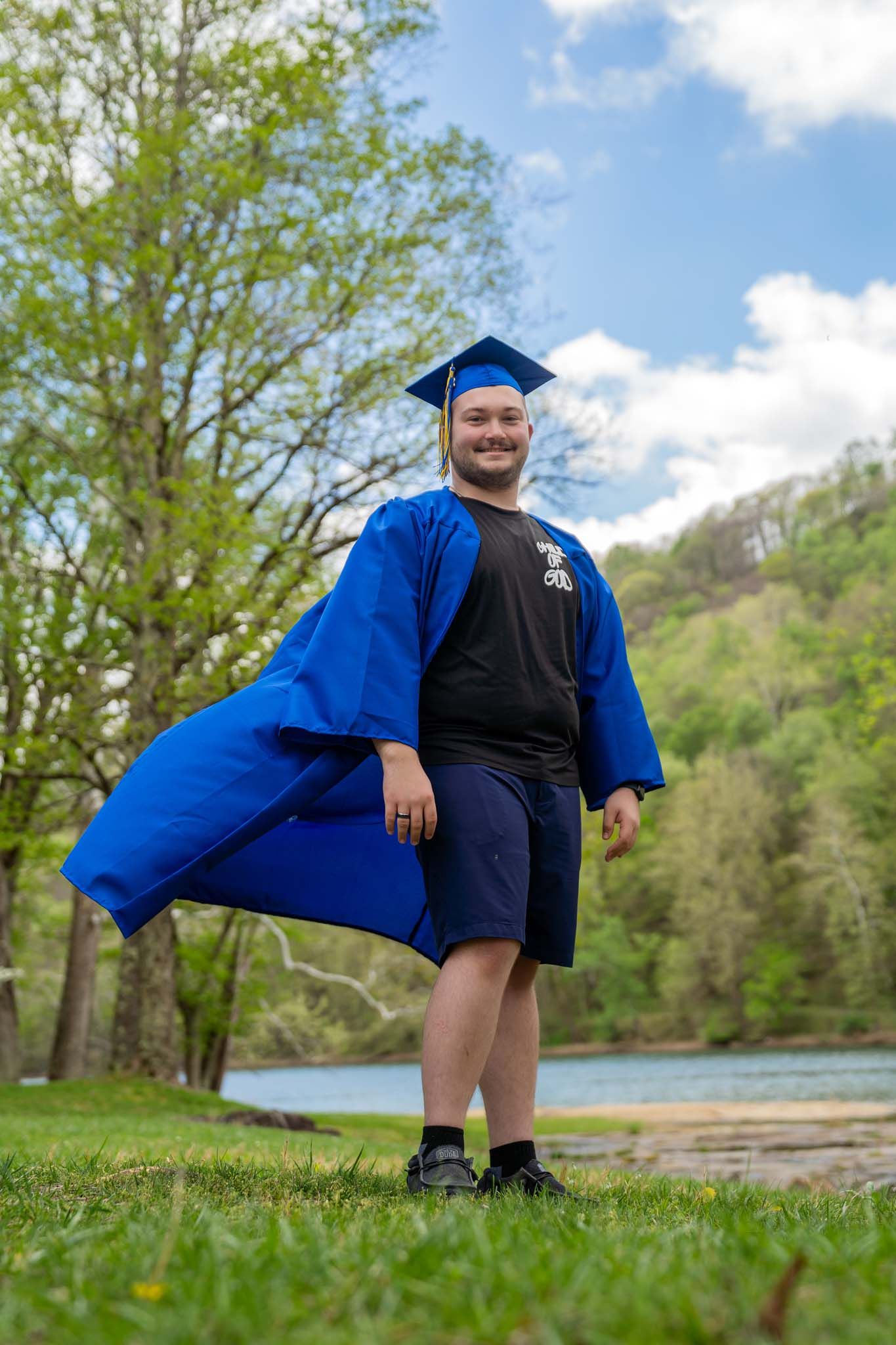 Smiling male graduate in blue cap and gown posing outdoors by river on sunny day.