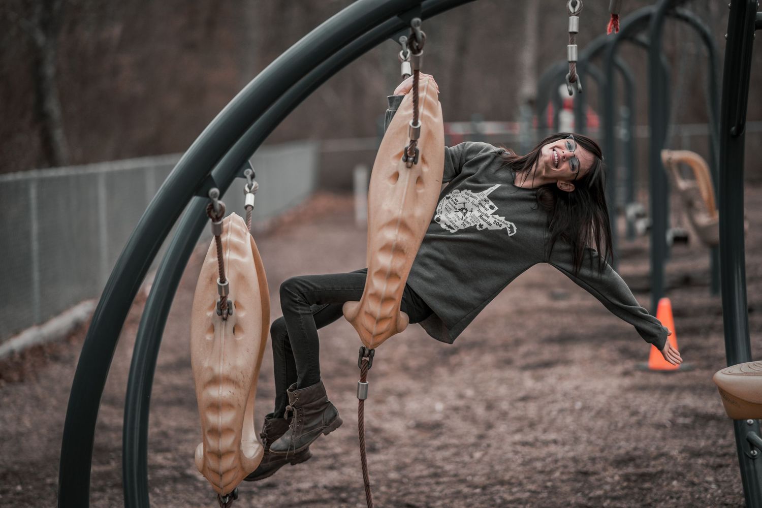 Person hanging sideways on outdoor gym equipment, smiling, wearing black hoodie and pants in a park setting.