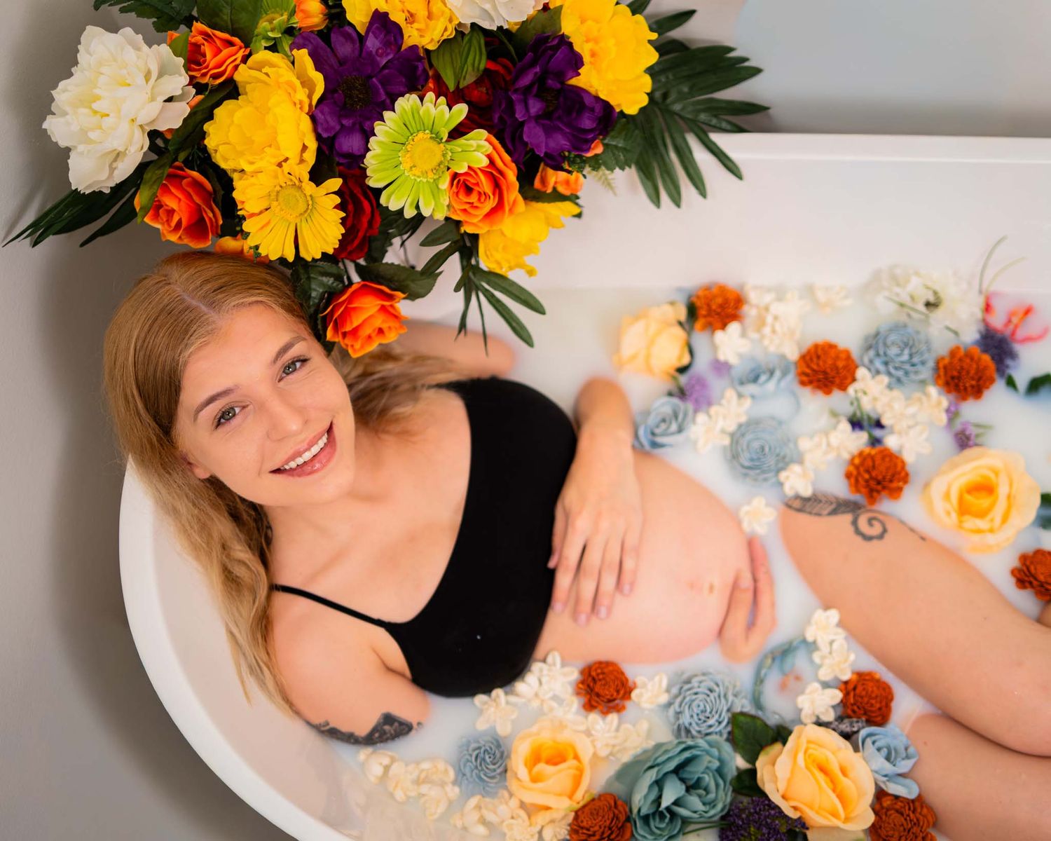Smiling pregnant woman in black bikini top lying in floral milk bath with colorful blooms.