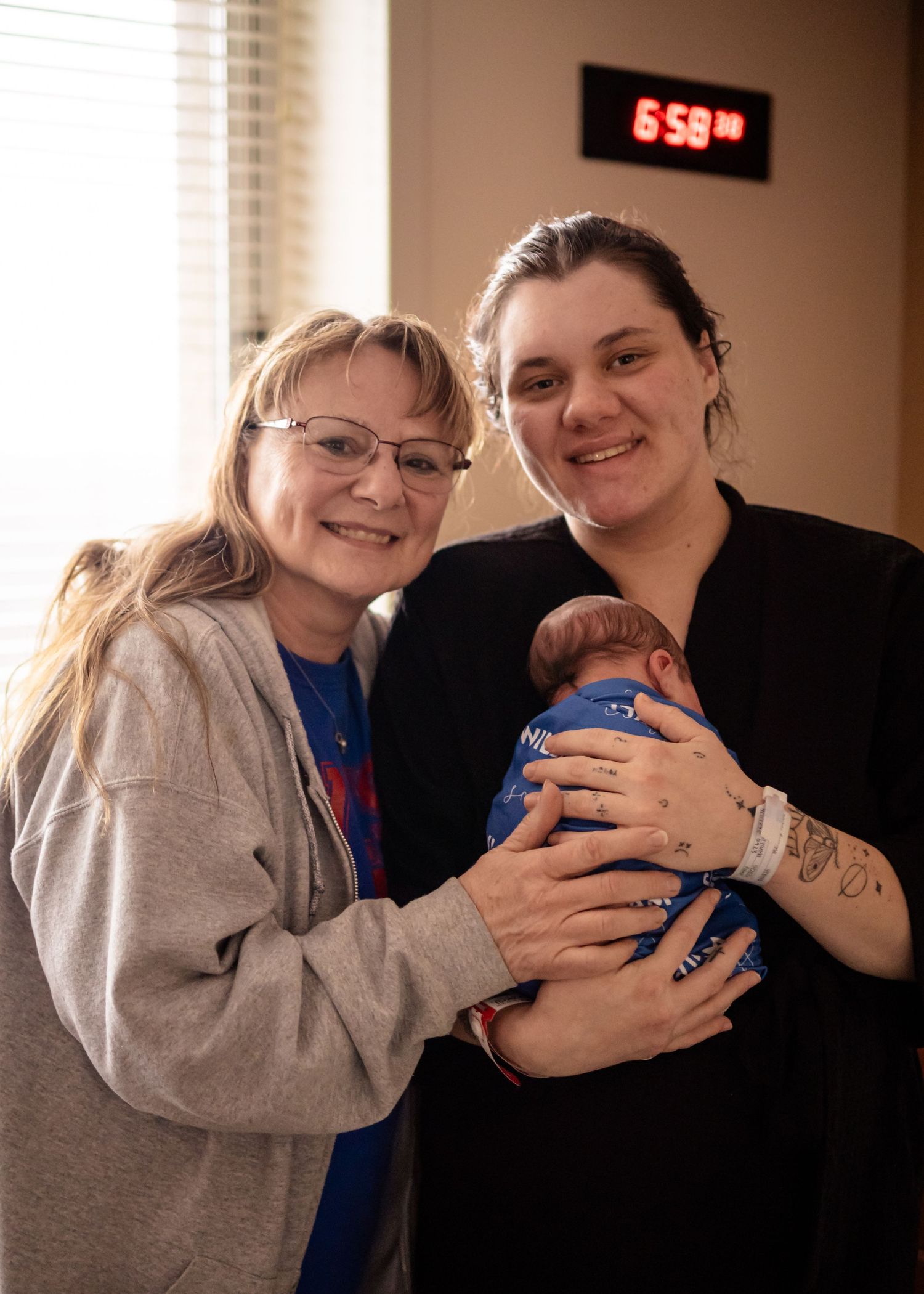 Two women smiling and holding a newborn baby in a blue outfit indoors near a window.