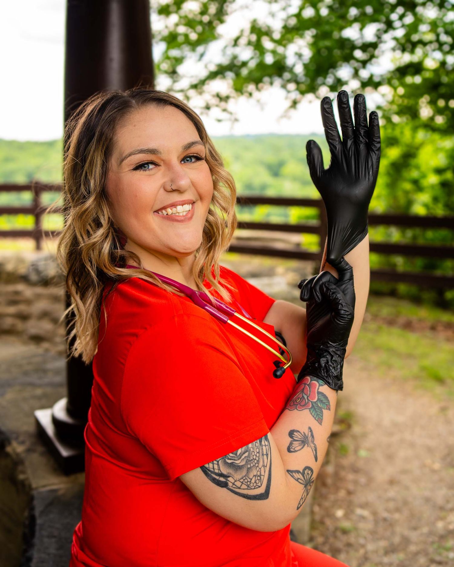 Smiling woman in red shirt putting on black gloves outdoors, showing tattoos on her arm with green trees in background.
