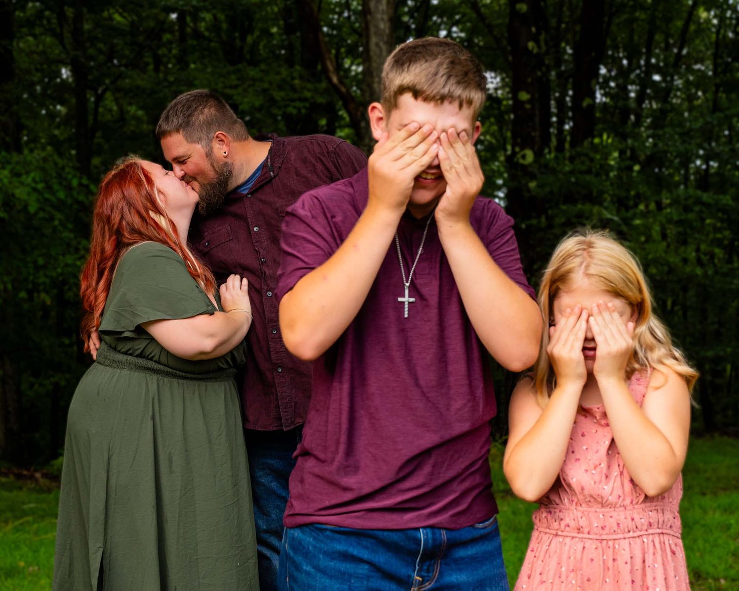Parents share a kiss behind their embarrassed teenage son and daughter covering their faces during a family photo session.