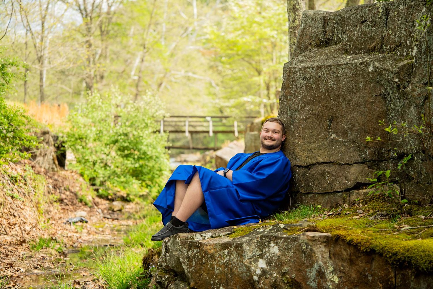 Female graduate in blue gown laughing while relaxing against a mossy stone wall in a forest setting.