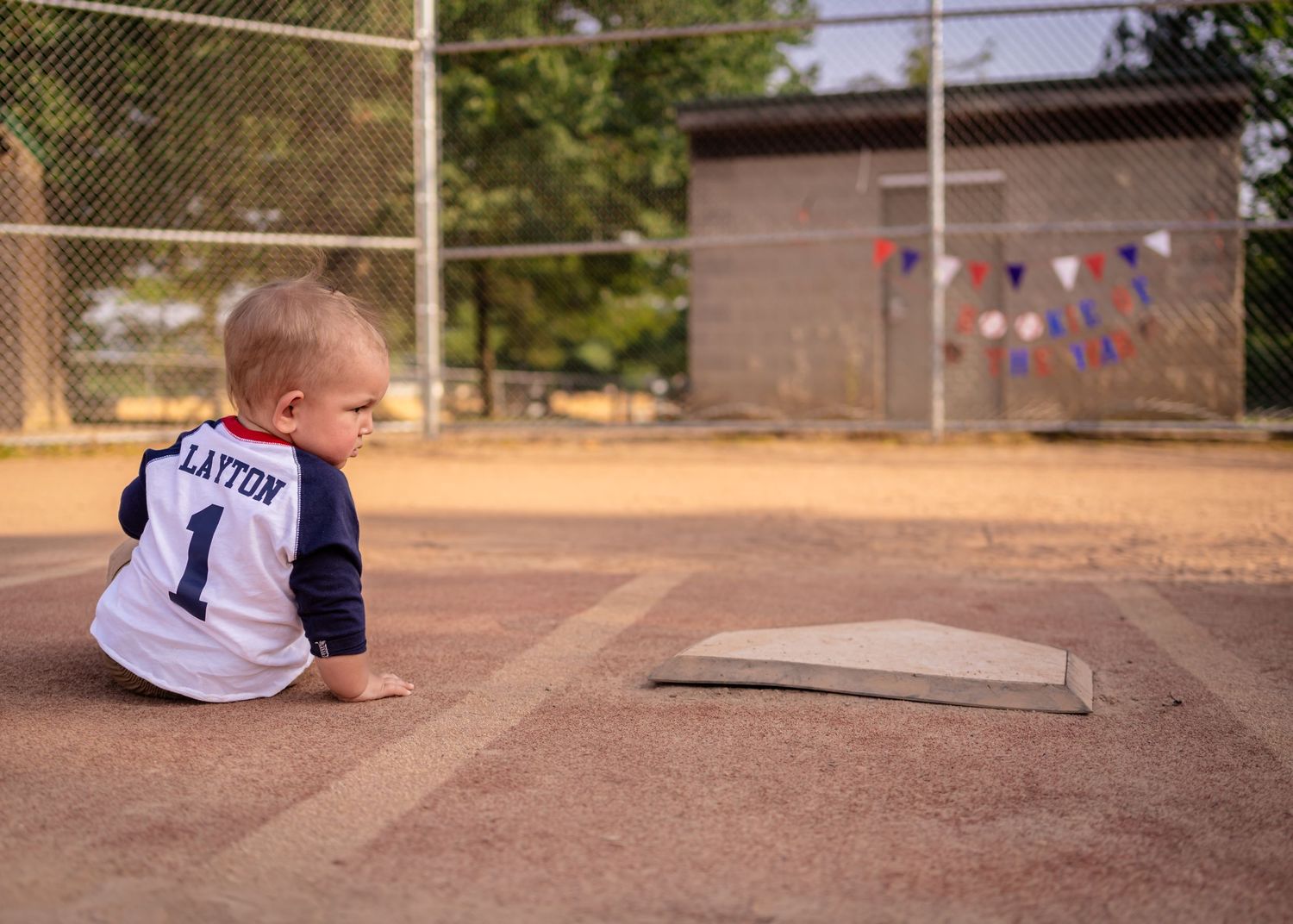 Baby in a baseball jersey sitting on a dirt field near home plate at a baseball diamond.