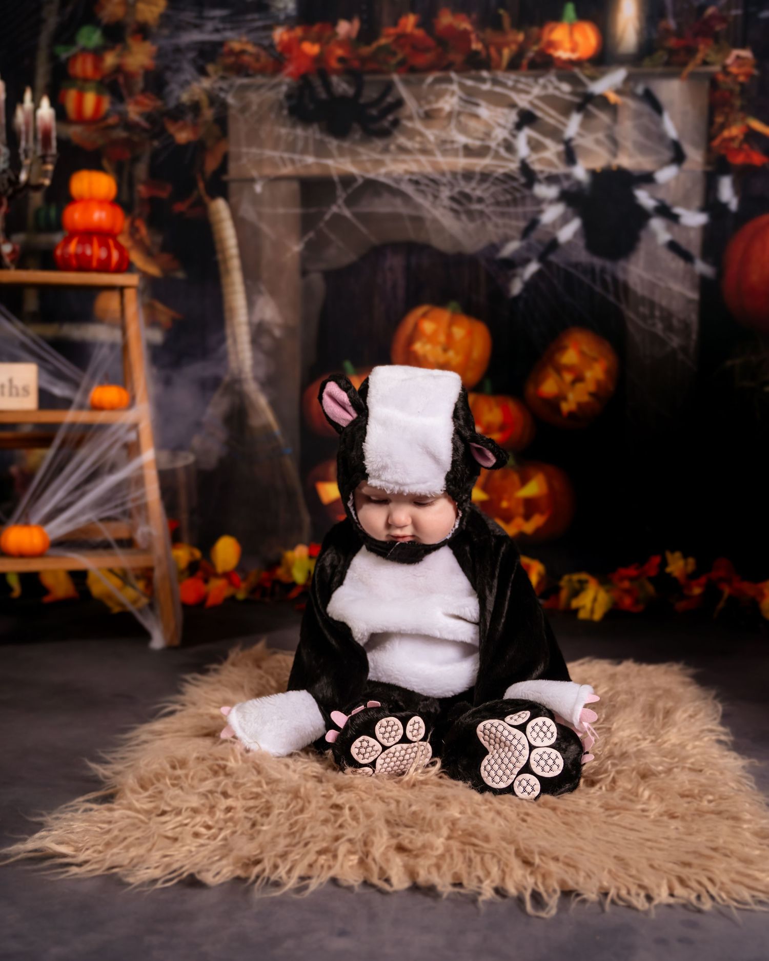 Baby in black and white skunk Halloween costume sits on fur rug surrounded by pumpkins and spooky decorations.