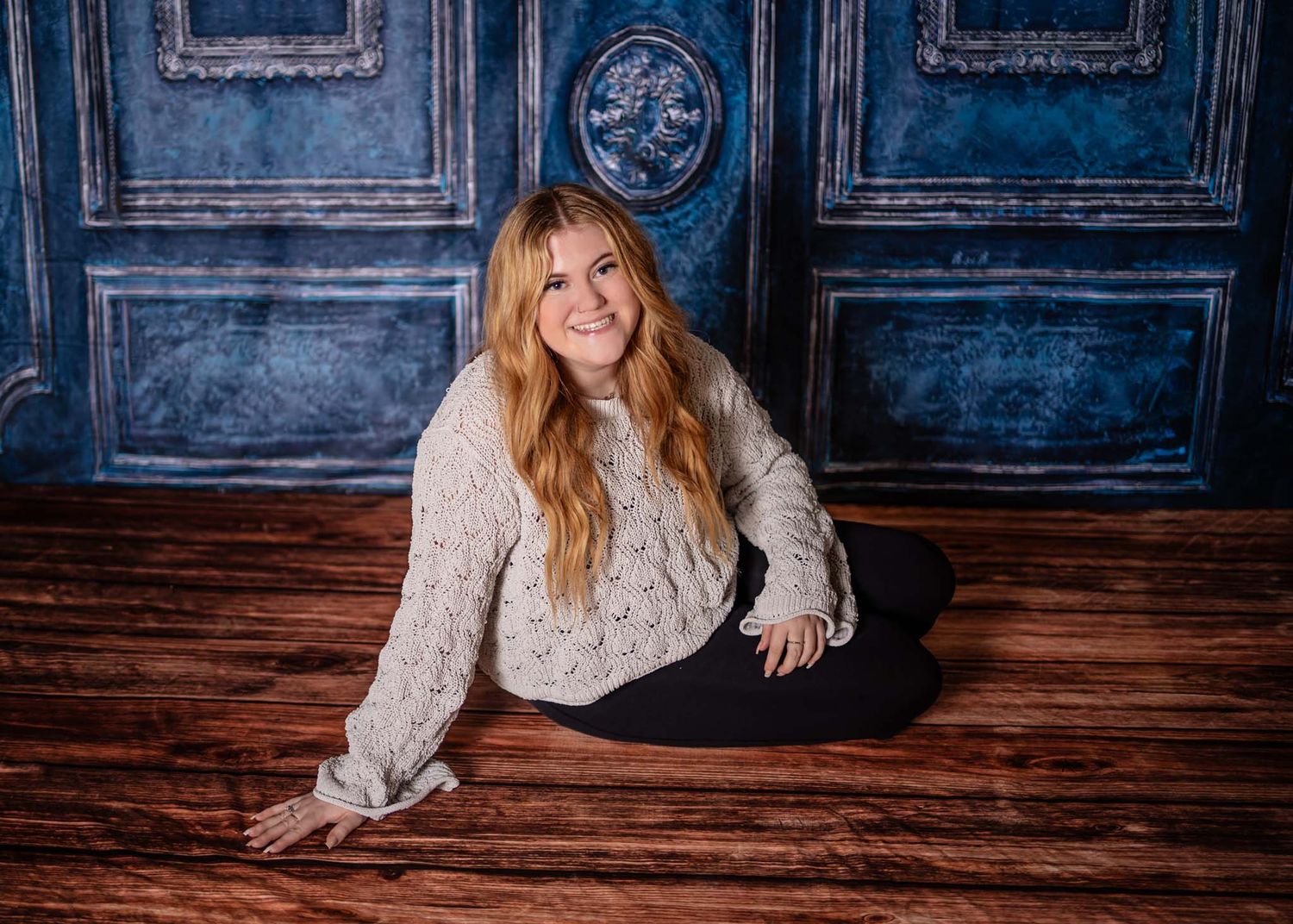 Young woman with long red hair sitting on floor smiling, blue vintage door backdrop.