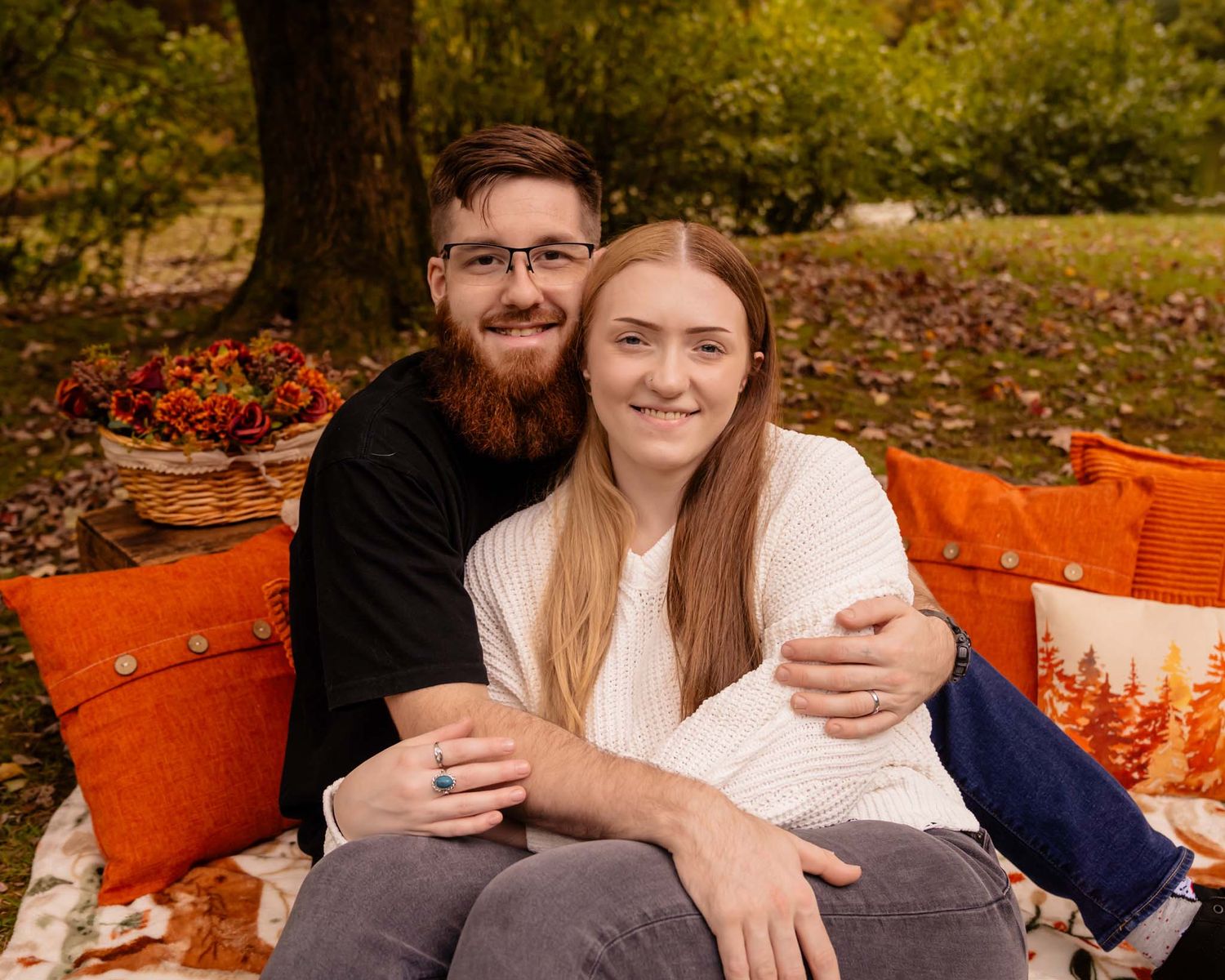 A happy couple sits together on a colorful autumn picnic blanket surrounded by fall foliage and orange decor.