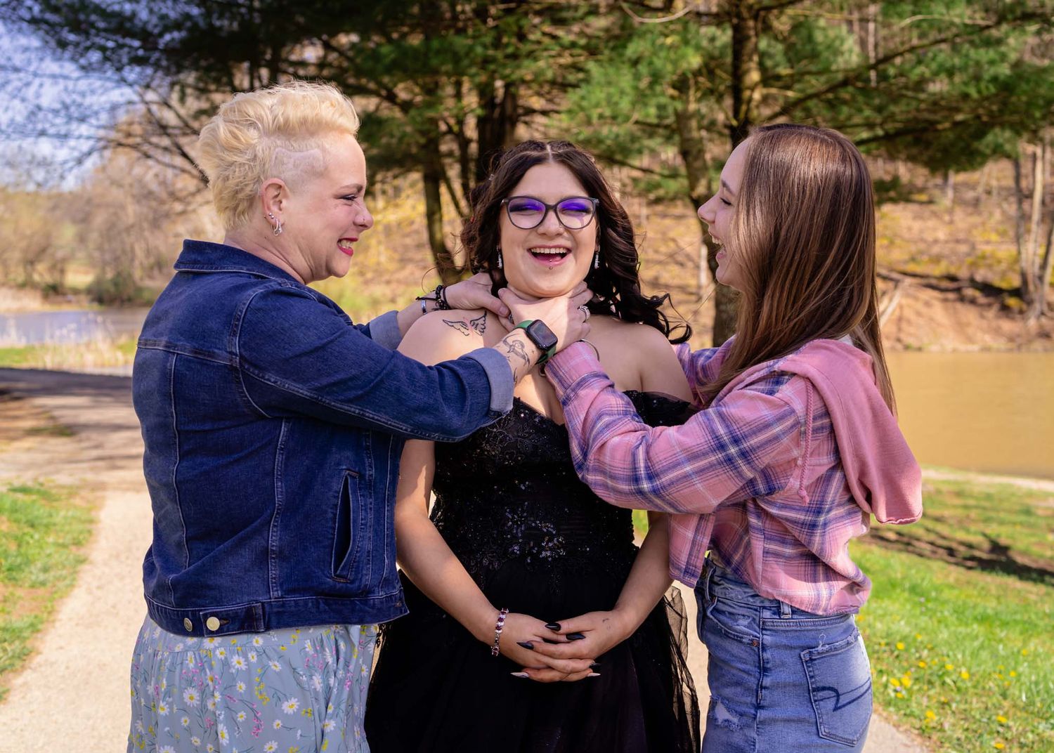 Three women laughing and interacting outdoors near a lake surrounded by spring trees.