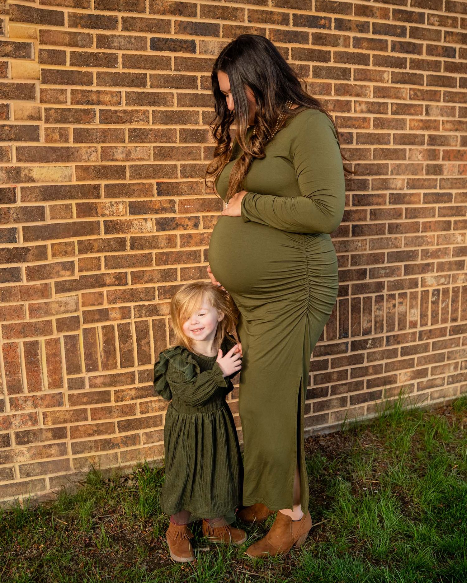 Pregnant woman in olive green dress poses with young daughter against brick wall for maternity photo.