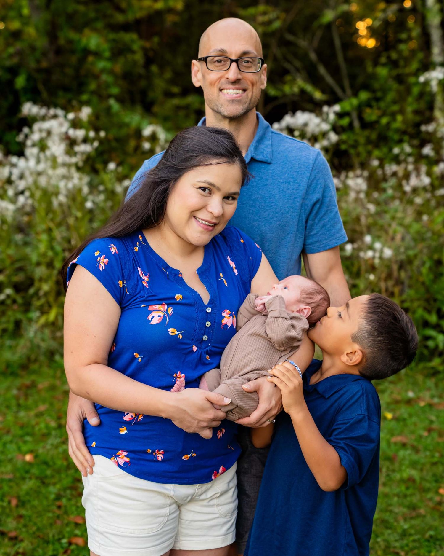 Smiling family of four outdoors, mother in blue floral top holding newborn while son kisses baby.