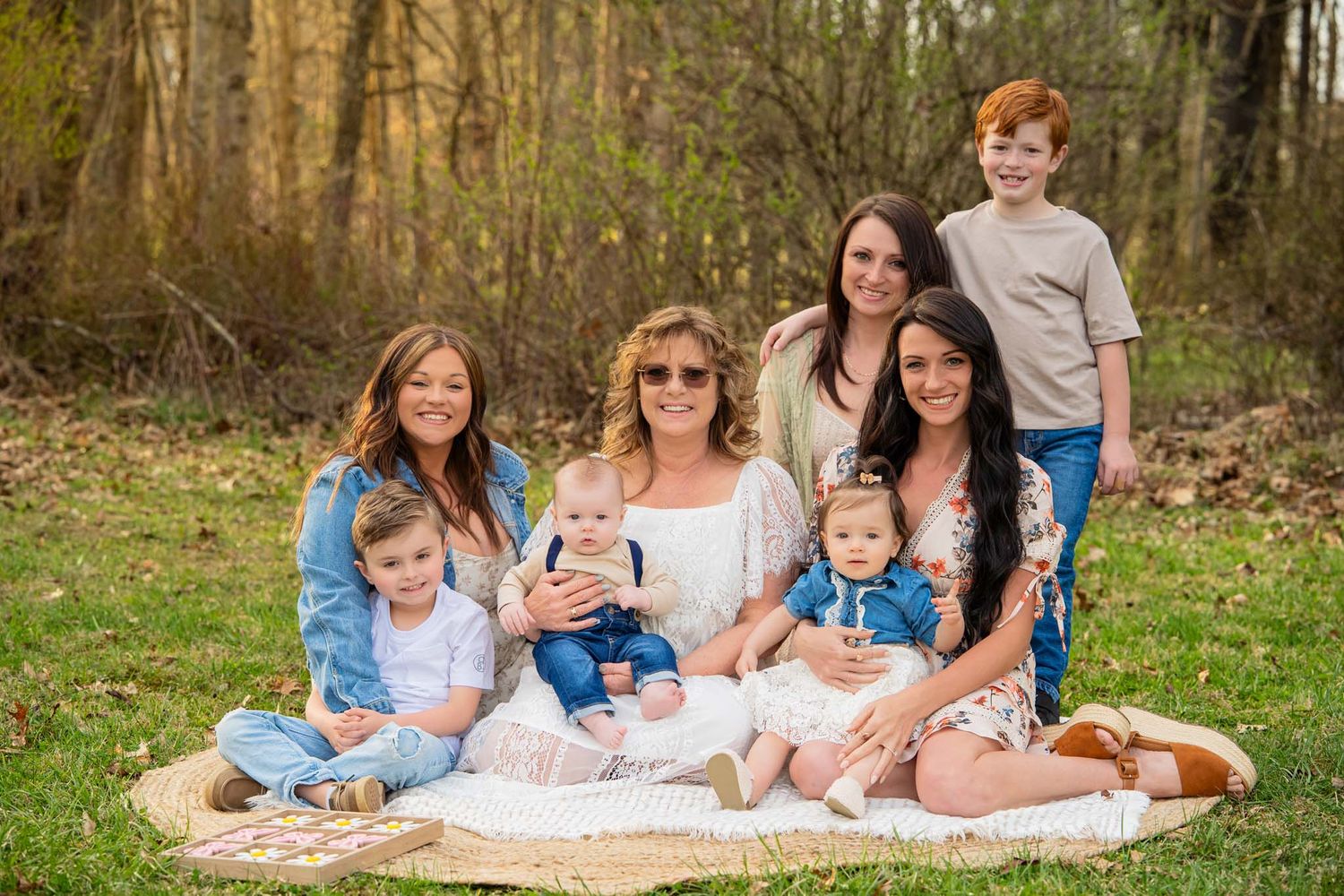 Family portrait with grandmother, three daughters, and four grandchildren sitting on a blanket outdoors in a wooded park.