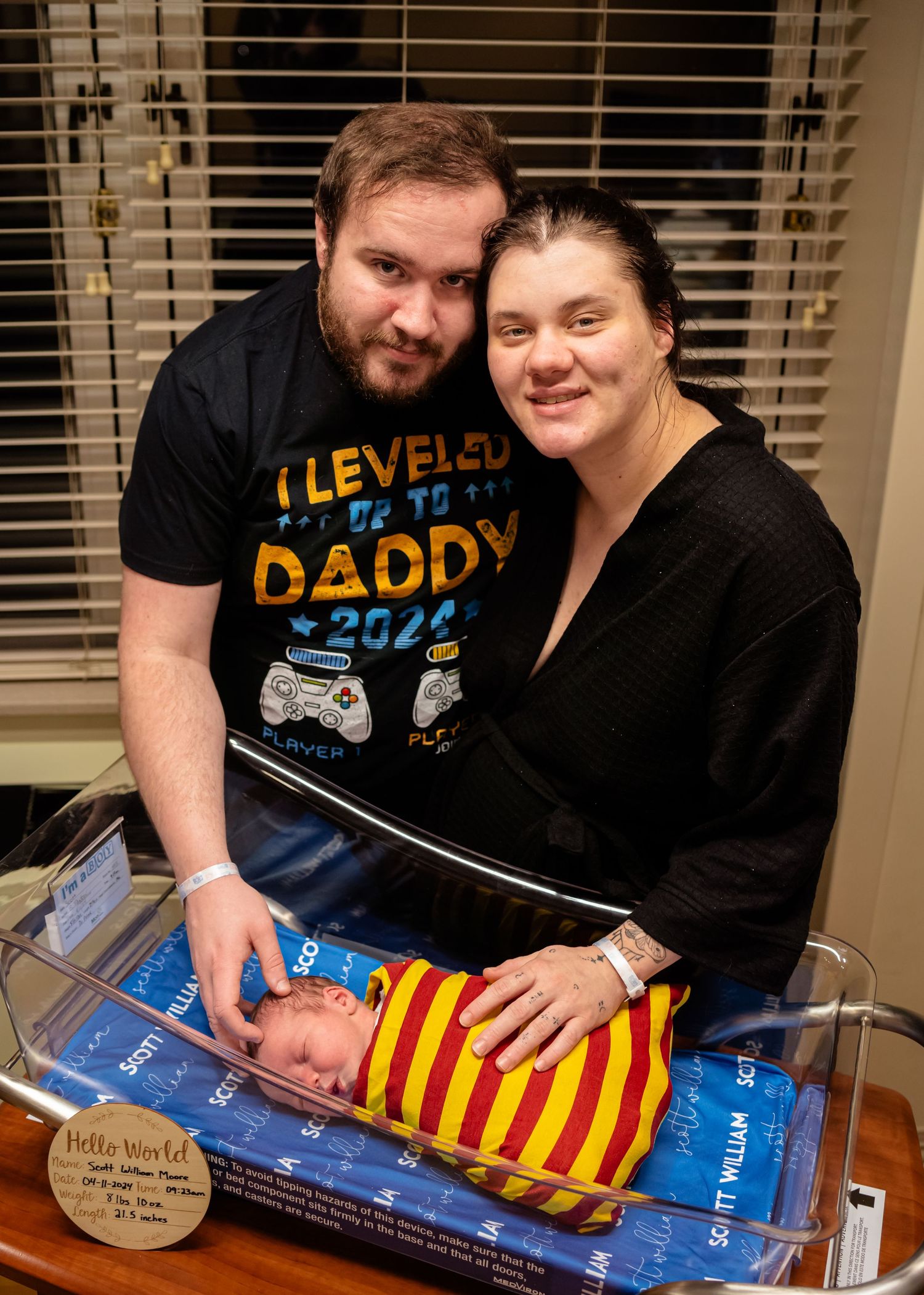 New parents smile proudly beside their newborn baby in a hospital bassinet, father wearing an I Leveled Up to Daddy shirt.