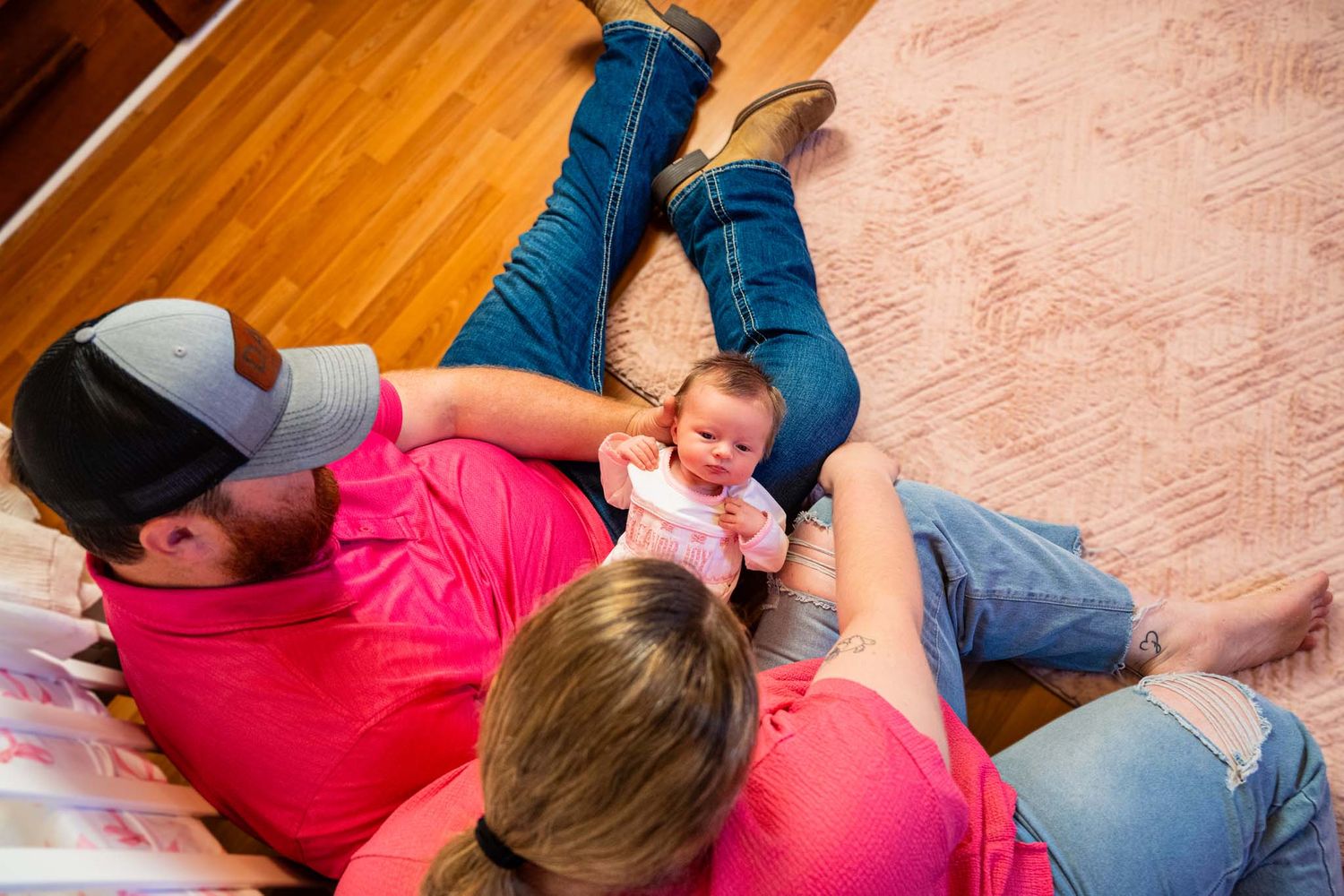 Two parents in pink shirts lying on hardwood floor bonding with their newborn baby at home.