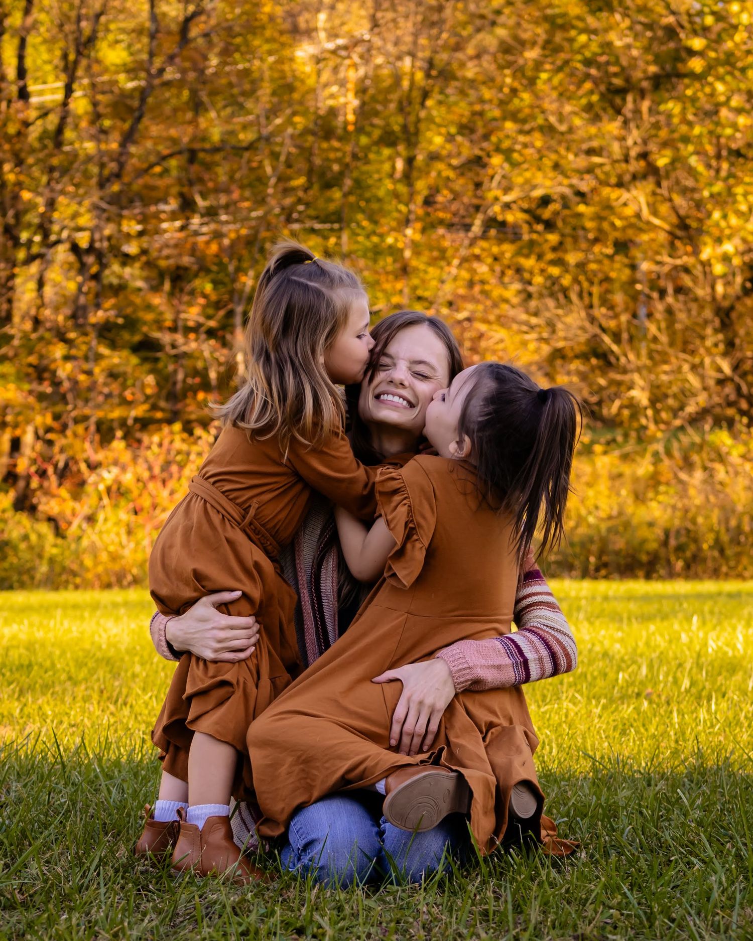 Joyful mother kneeling on grass as two young daughters in rust dresses give her kisses on the cheeks outdoors.