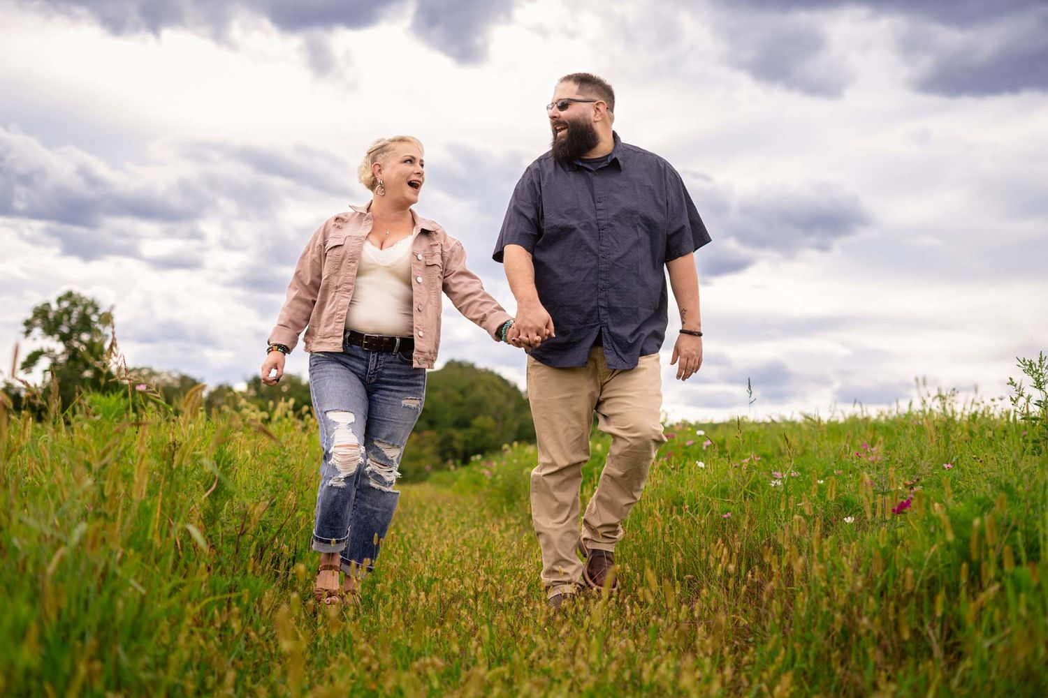 Happy couple walks hand in hand through green meadow under dramatic stormy sky.