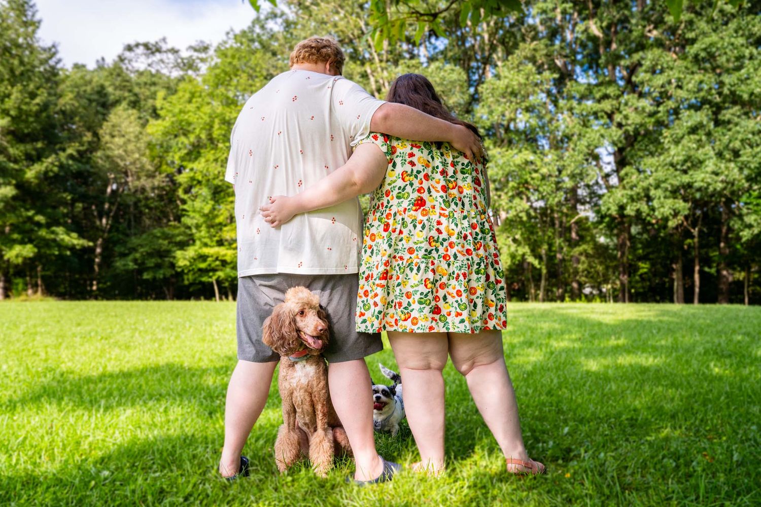 Couple stands back to back embracing in green park with fluffy brown poodle sitting between their feet.