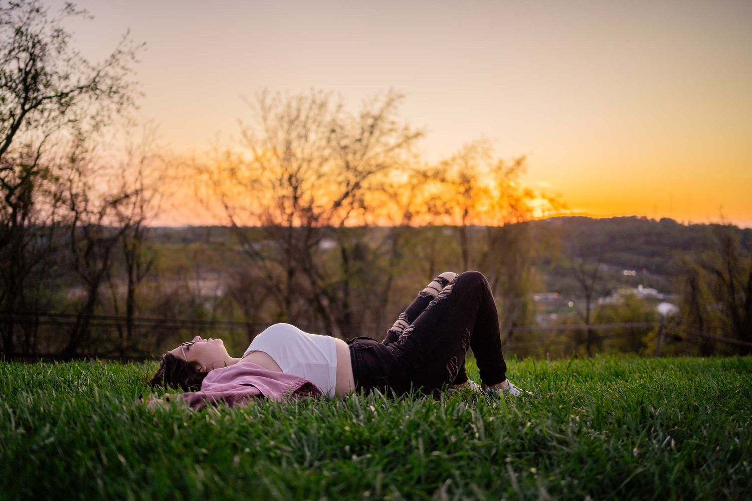 A young woman lies on green grass during a golden sunset in a serene outdoor setting.