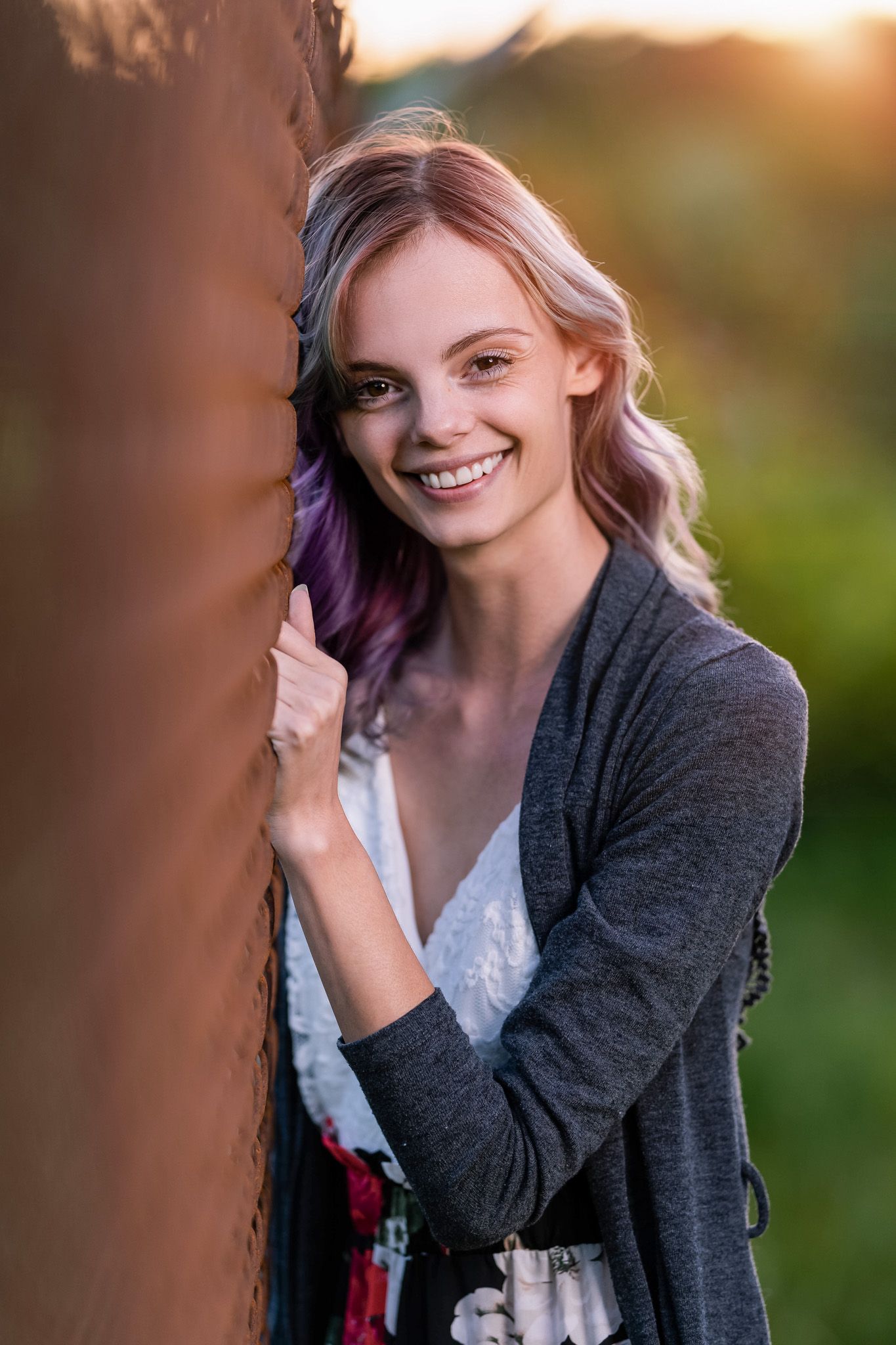 Smiling young woman with purple-tinted hair leaning against a wall at golden hour.