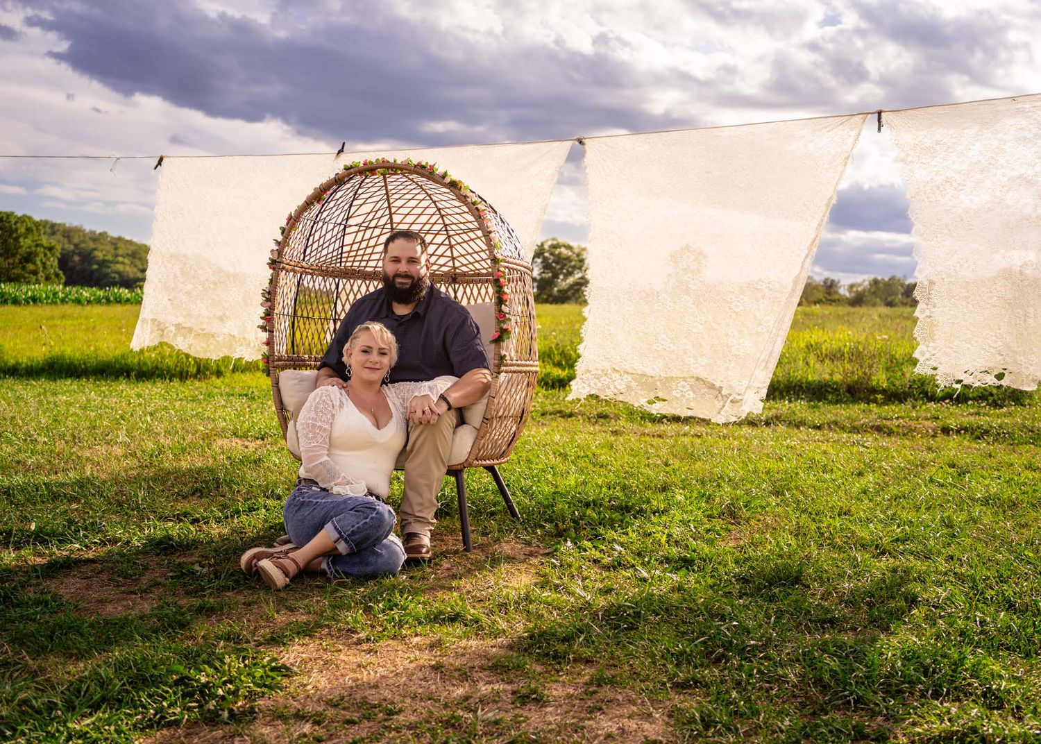 Couple poses together in rustic outdoor setting with wicker chair and hanging linens.