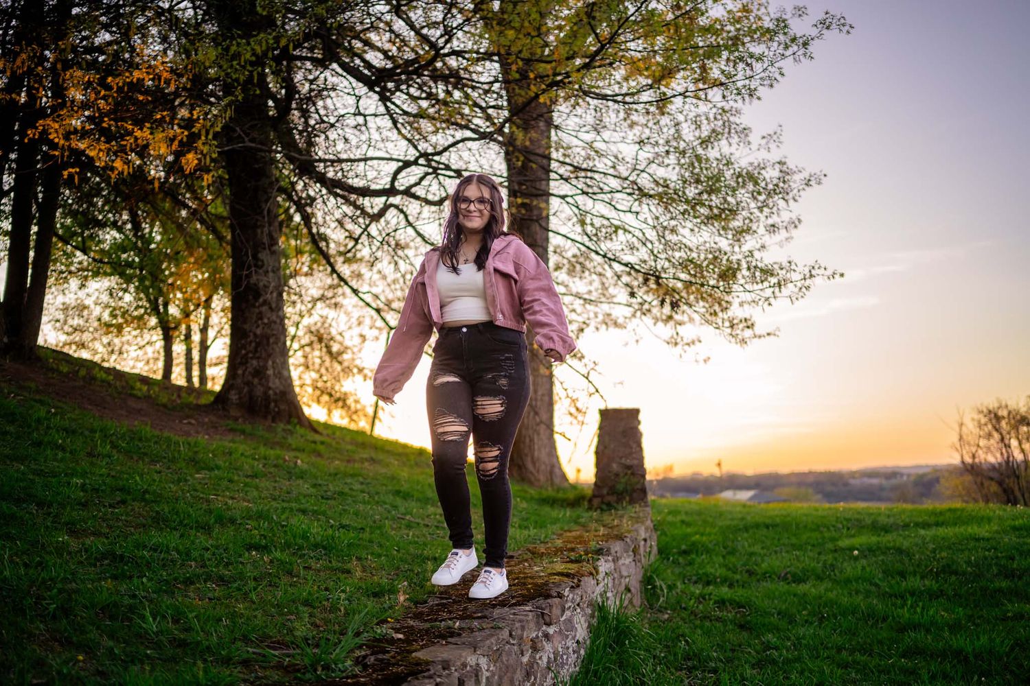 A smiling girl in a pink jacket and ripped jeans stands on a stone wall at sunset.