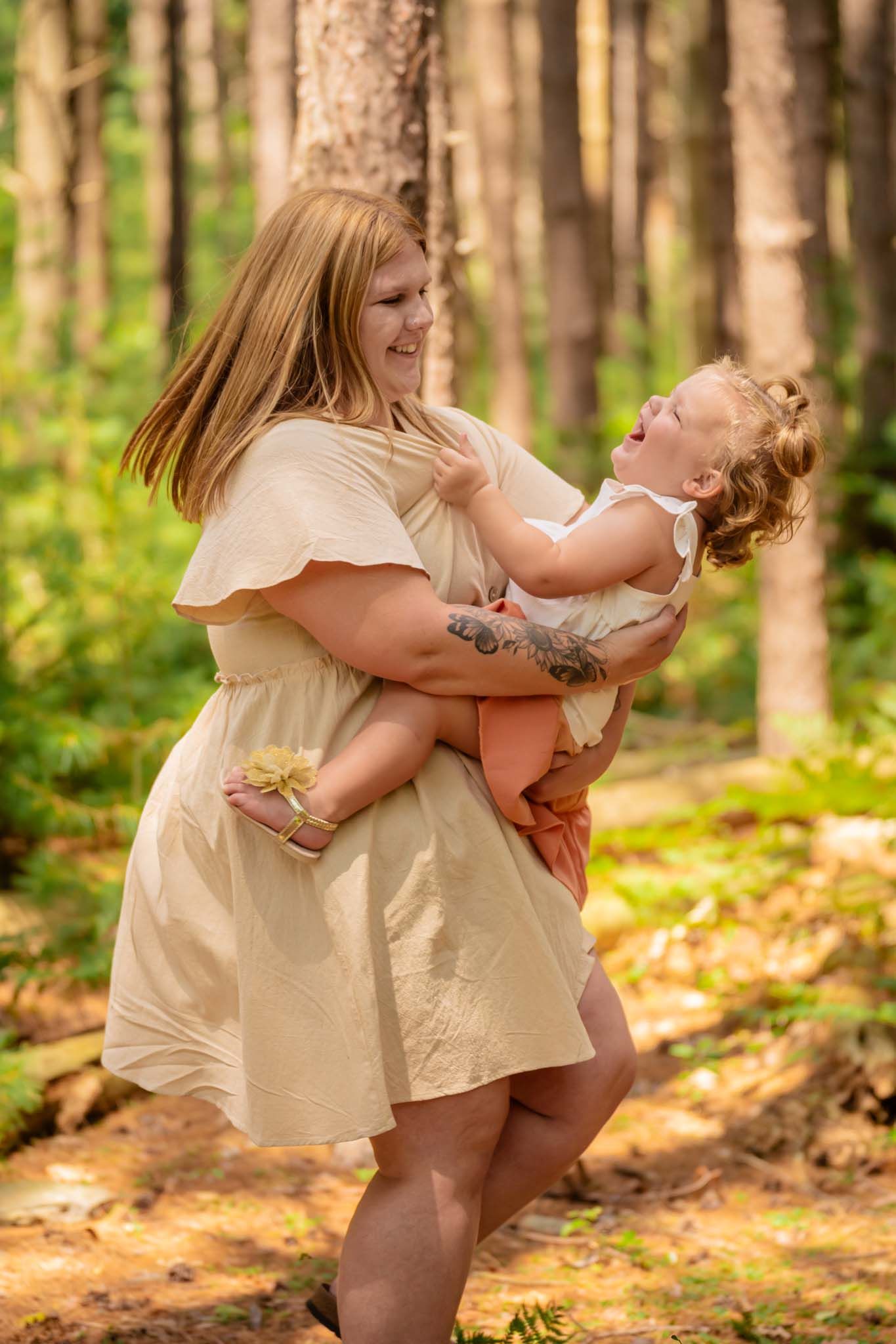 Mother laughing while holding joyful toddler daughter in sunny forest.