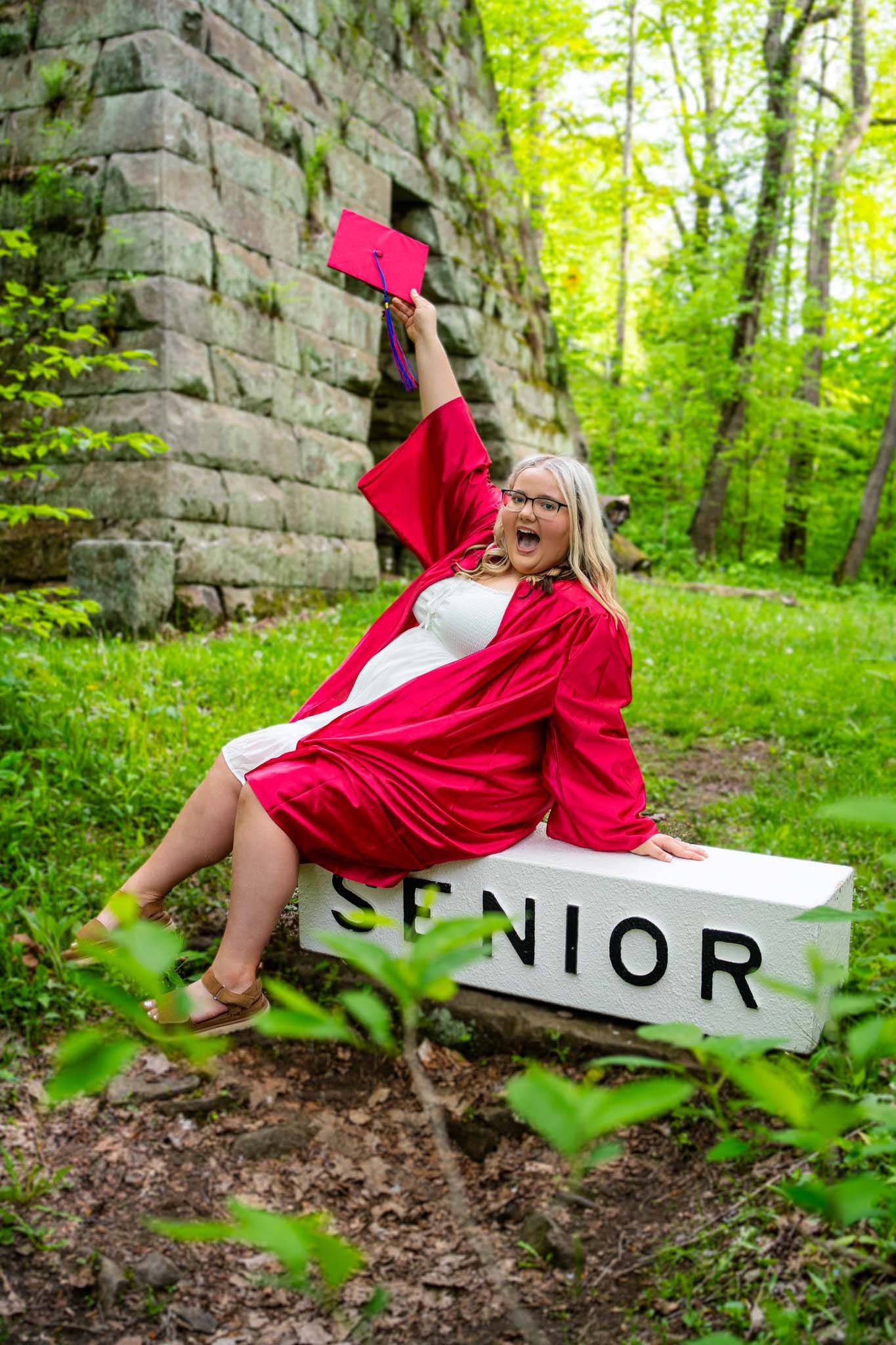 Excited graduate in red cap and gown sits on Senior bench outdoors.