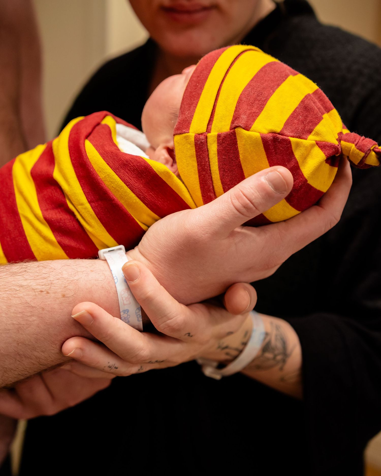 Newborn baby in red and yellow striped swaddle blanket and hat held by parents wearing hospital wristbands.
