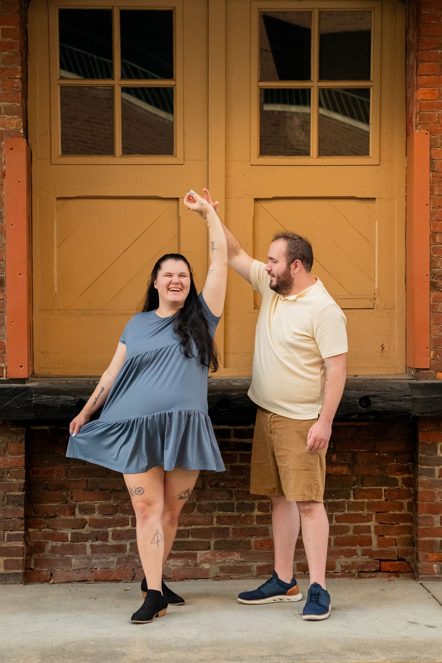 A couple dances outdoors, man in yellow polo twirling woman in blue dress before brick wall with yellow doors.