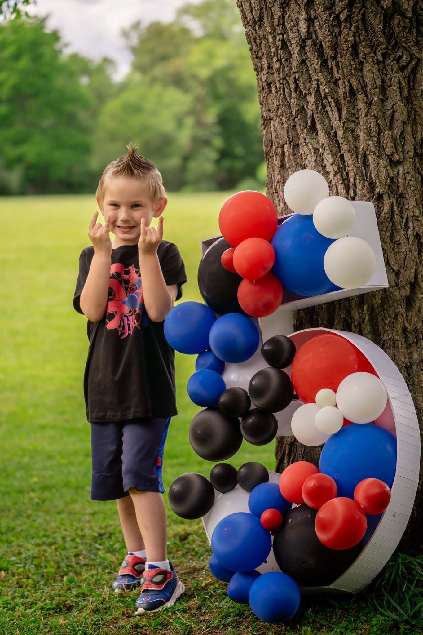 Young boy posing next to a large number 5 decorated with colorful balloons by a tree at an outdoor birthday party.