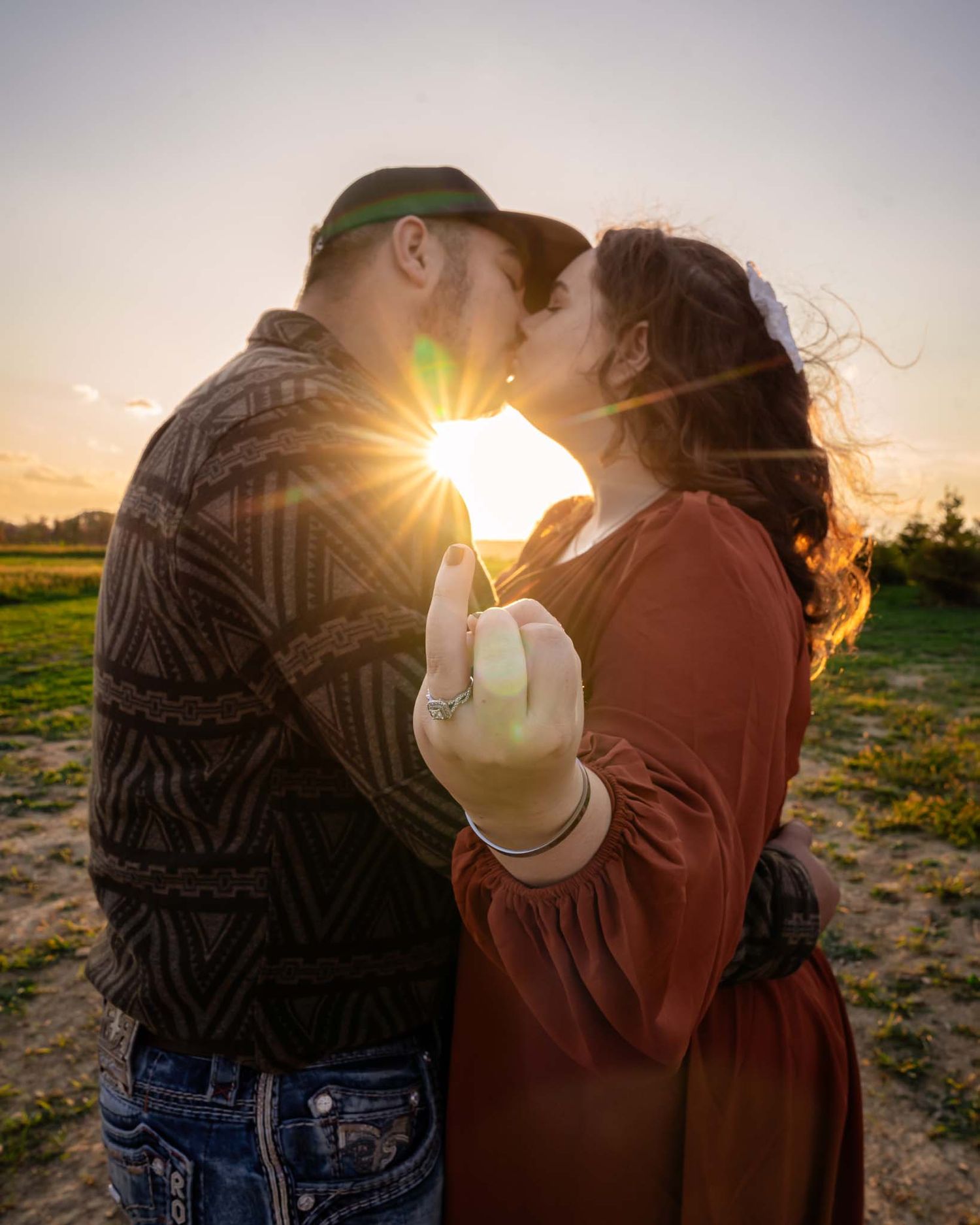 Couple sharing a romantic sunset kiss outdoors with golden sun flare and engagement ring visible.