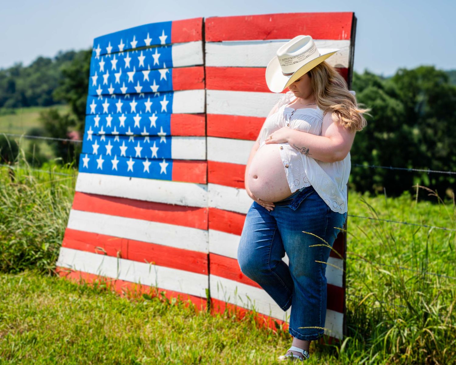 Pregnant woman in cowboy hat and jeans posing against a rustic wooden American flag sign in a green field.