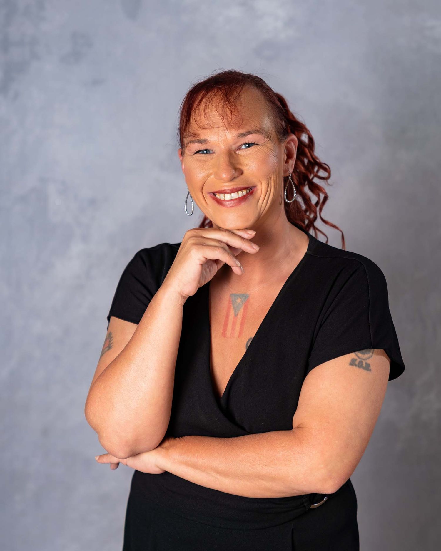 Smiling redhead woman in black wrap top posing with hand on chin against gray studio background.