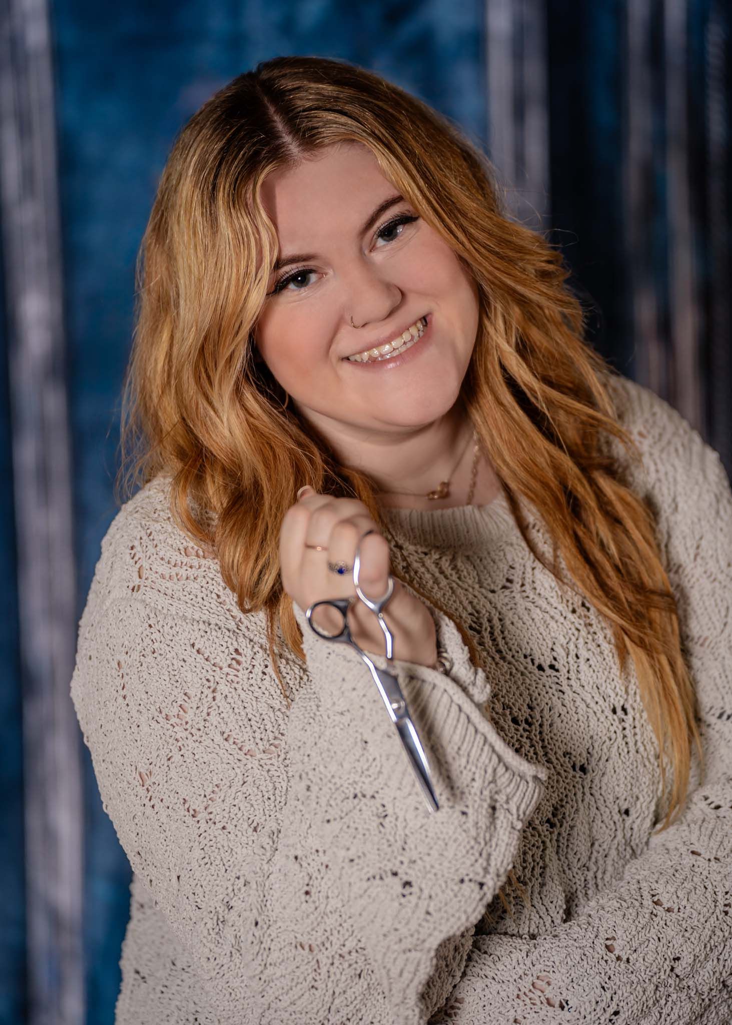 Smiling young woman with red hair holding scissors in cream knit sweater, blue backdrop.