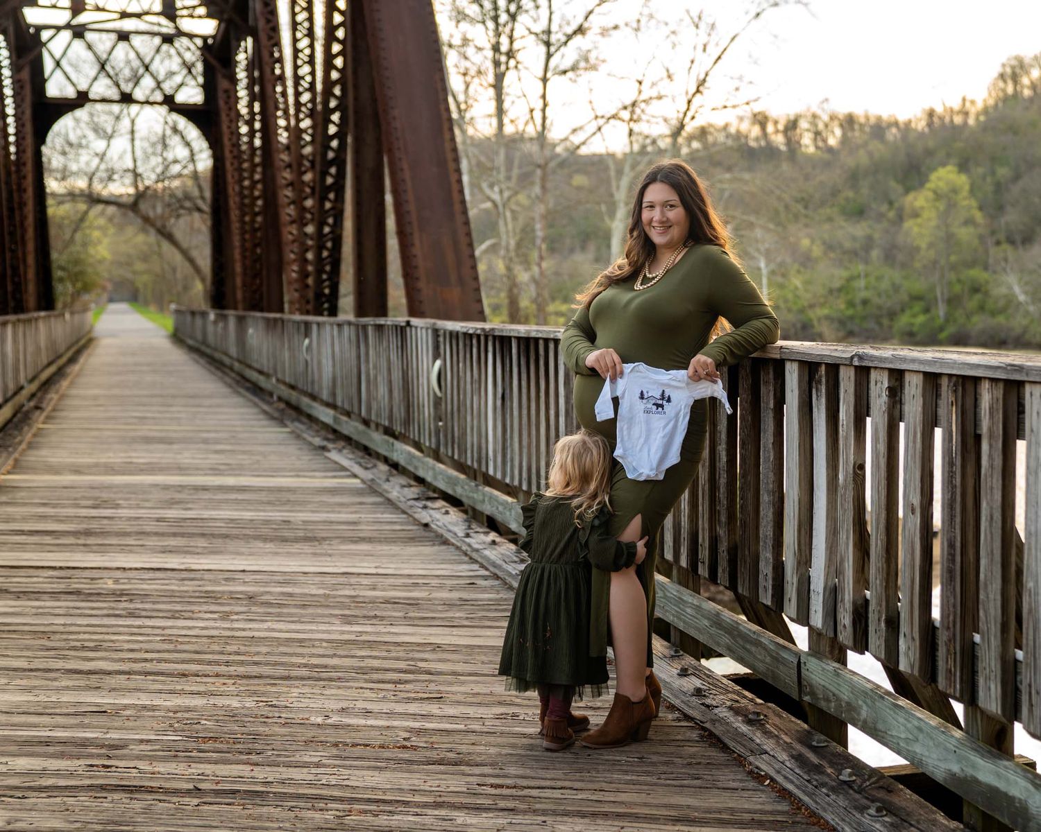A pregnant woman in green holds a baby onesie on a wooden bridge with a toddler nearby.
